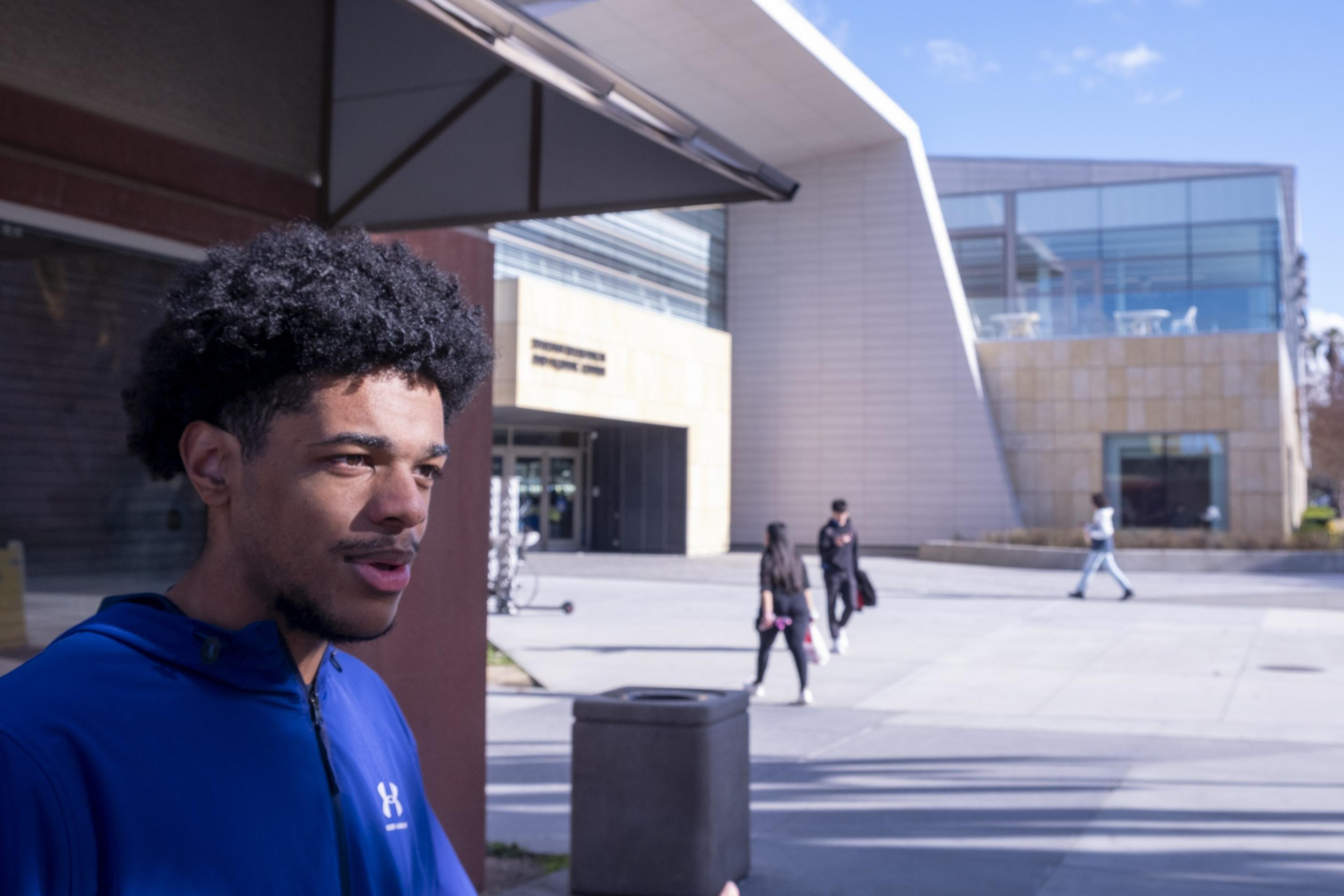 A person in a blue jacket is standing outdoors near a modern building with large windows. Other people walk by in the background under a sunny sky.