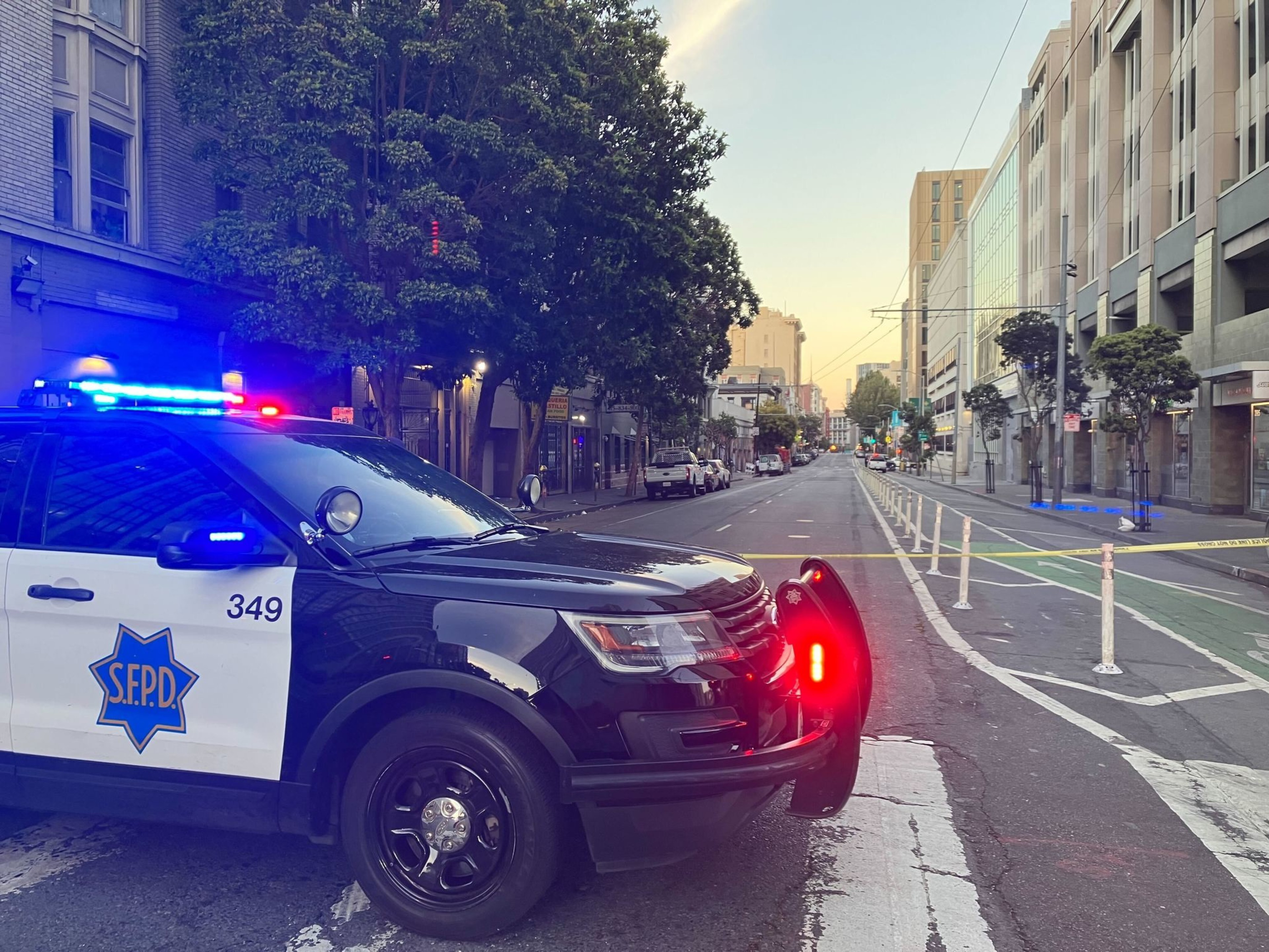 A police SUV with flashing lights blocking a city street at dawn or dusk, with yellow tape suggesting a closed-off area.