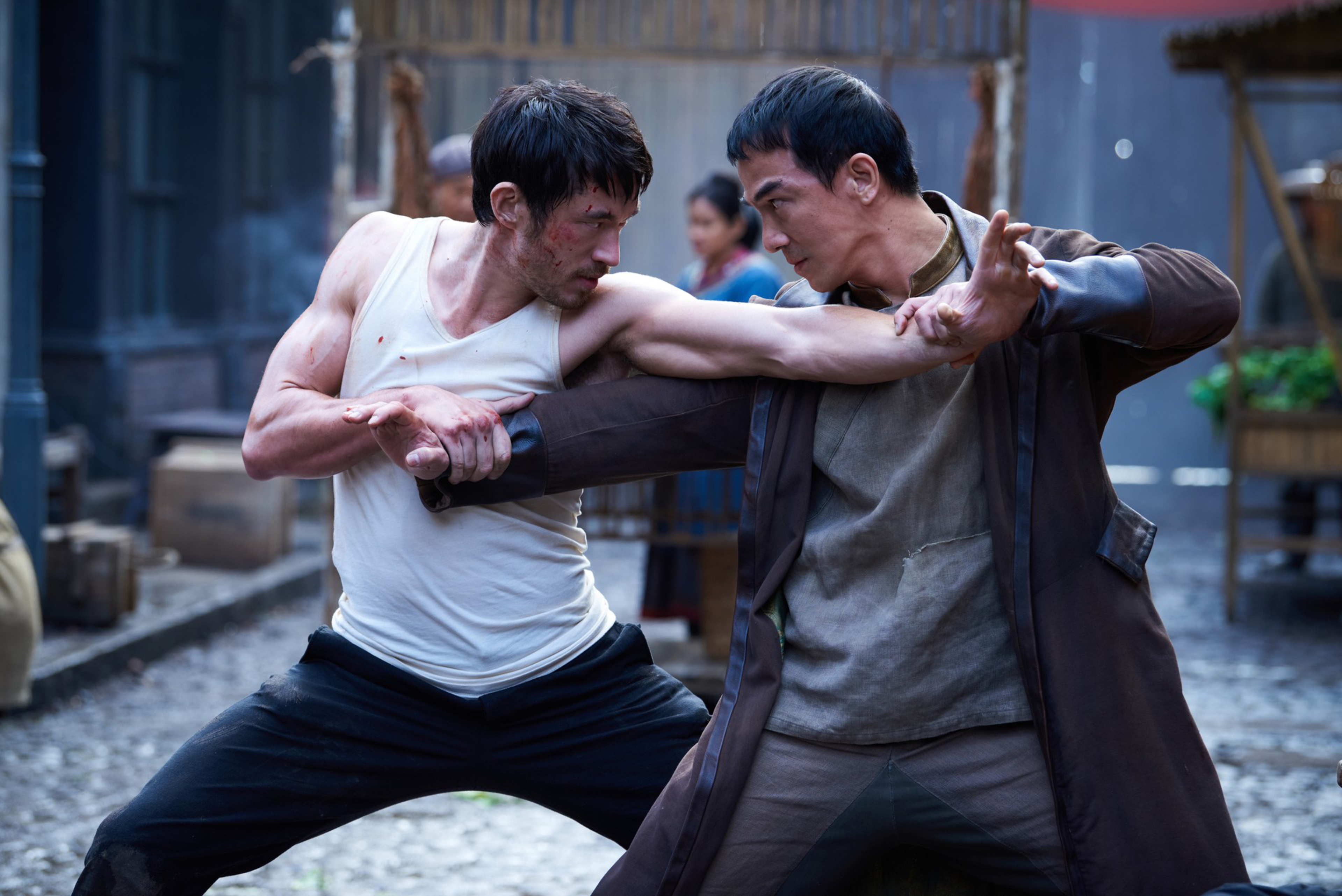 Two men in intense martial arts combat, one in a white tank top with bruises, the other in a dark coat, gripping each other's arms.
