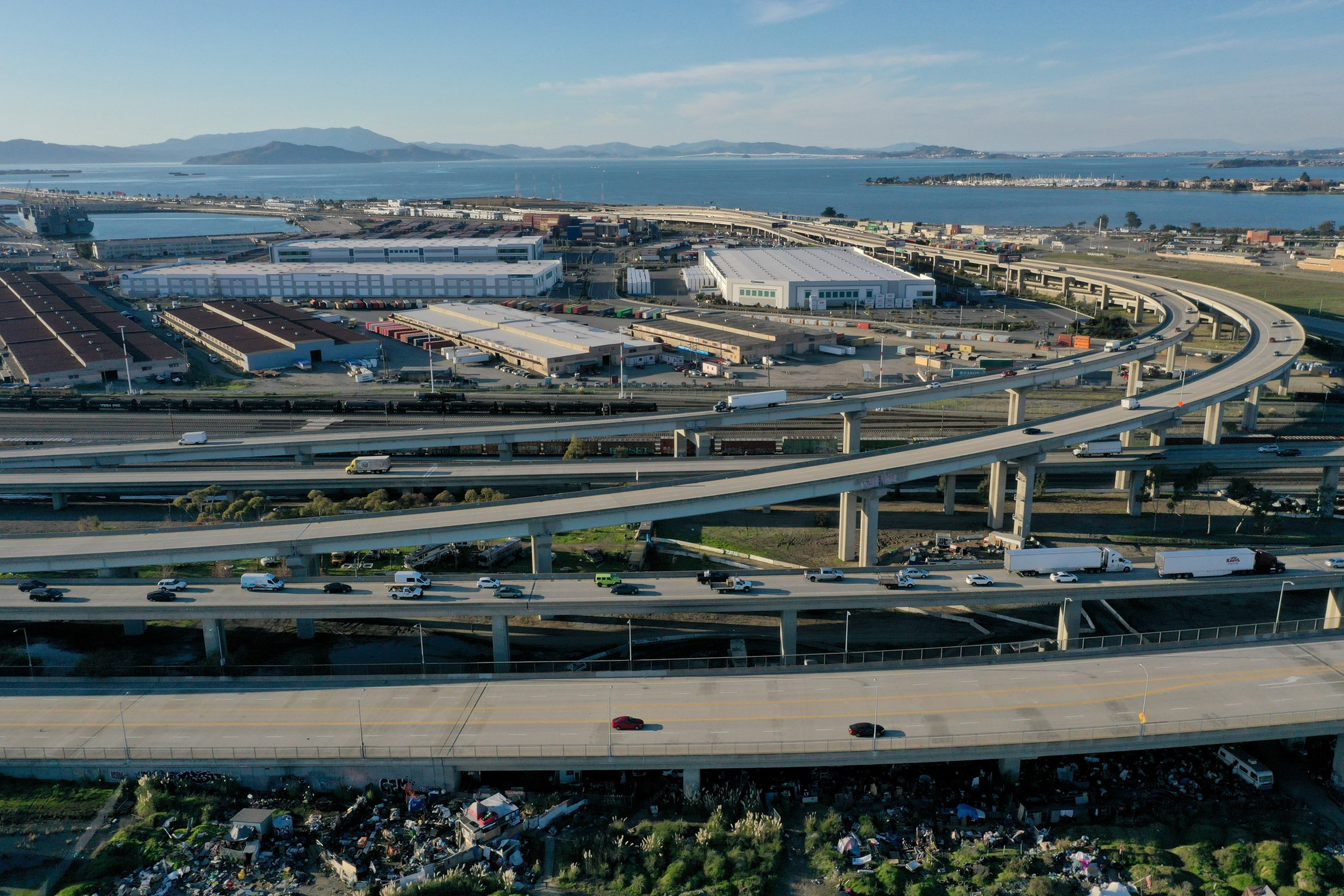 Multiple layered highways curve over an industrial area near water, with vehicles traveling on them and mountains visible in the distance under a clear sky.