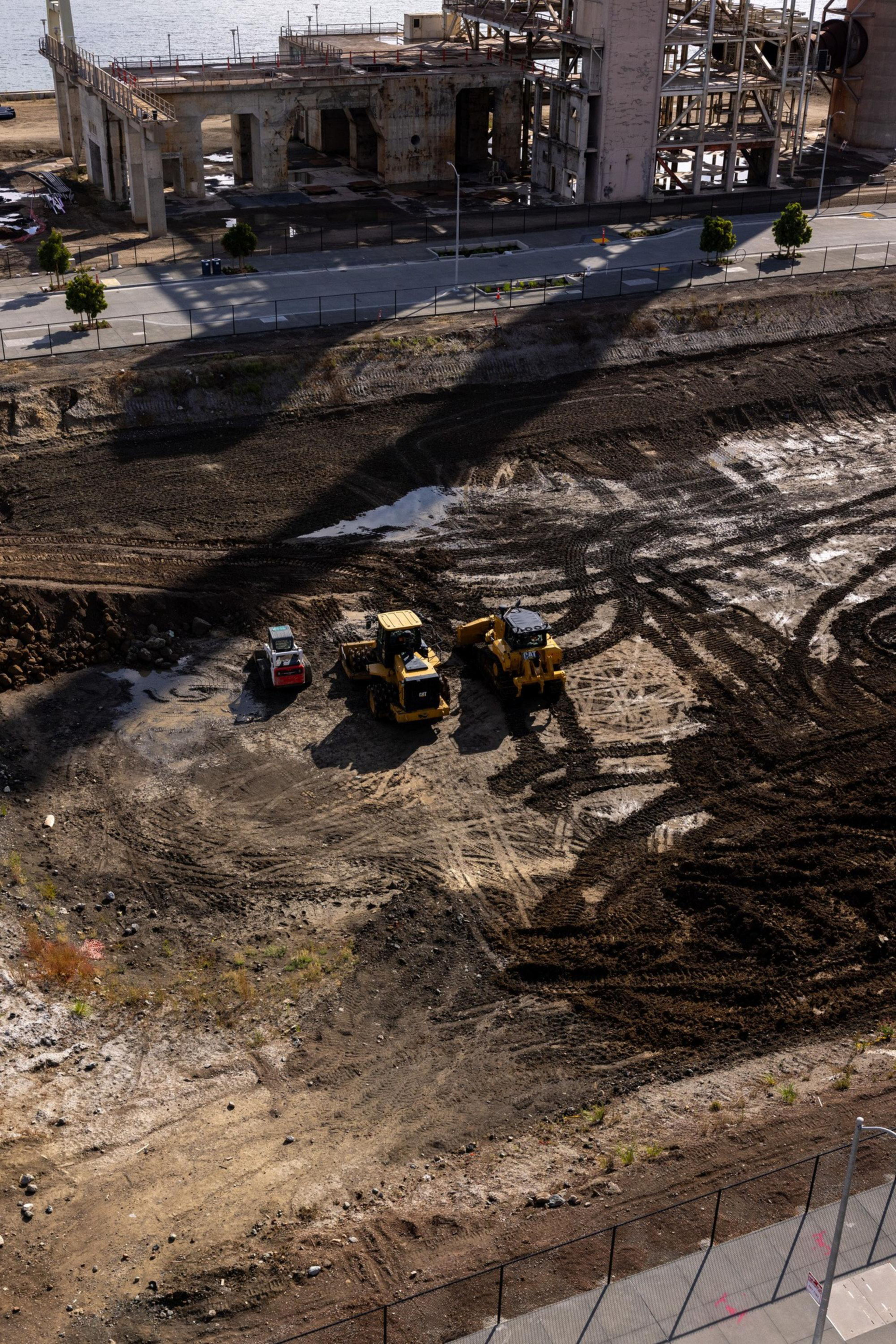Three construction vehicles are parked on a muddy, tracked dirt lot near a fenced industrial area with a partially demolished building.