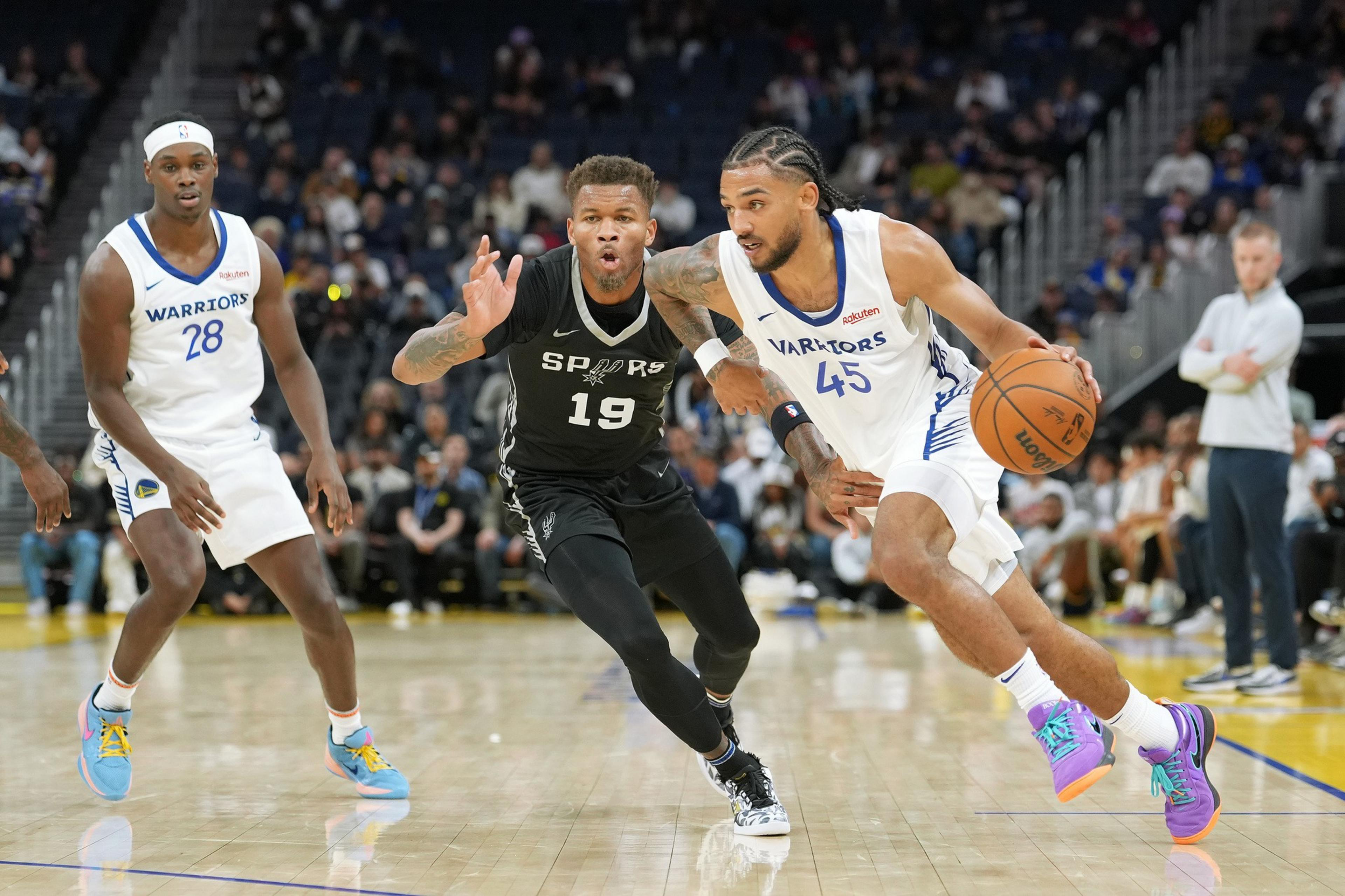 A basketball player in a white &quot;Warriors&quot; jersey drives forward with the ball, closely followed by a defender in a black &quot;Spurs&quot; jersey. Another player in white watches.