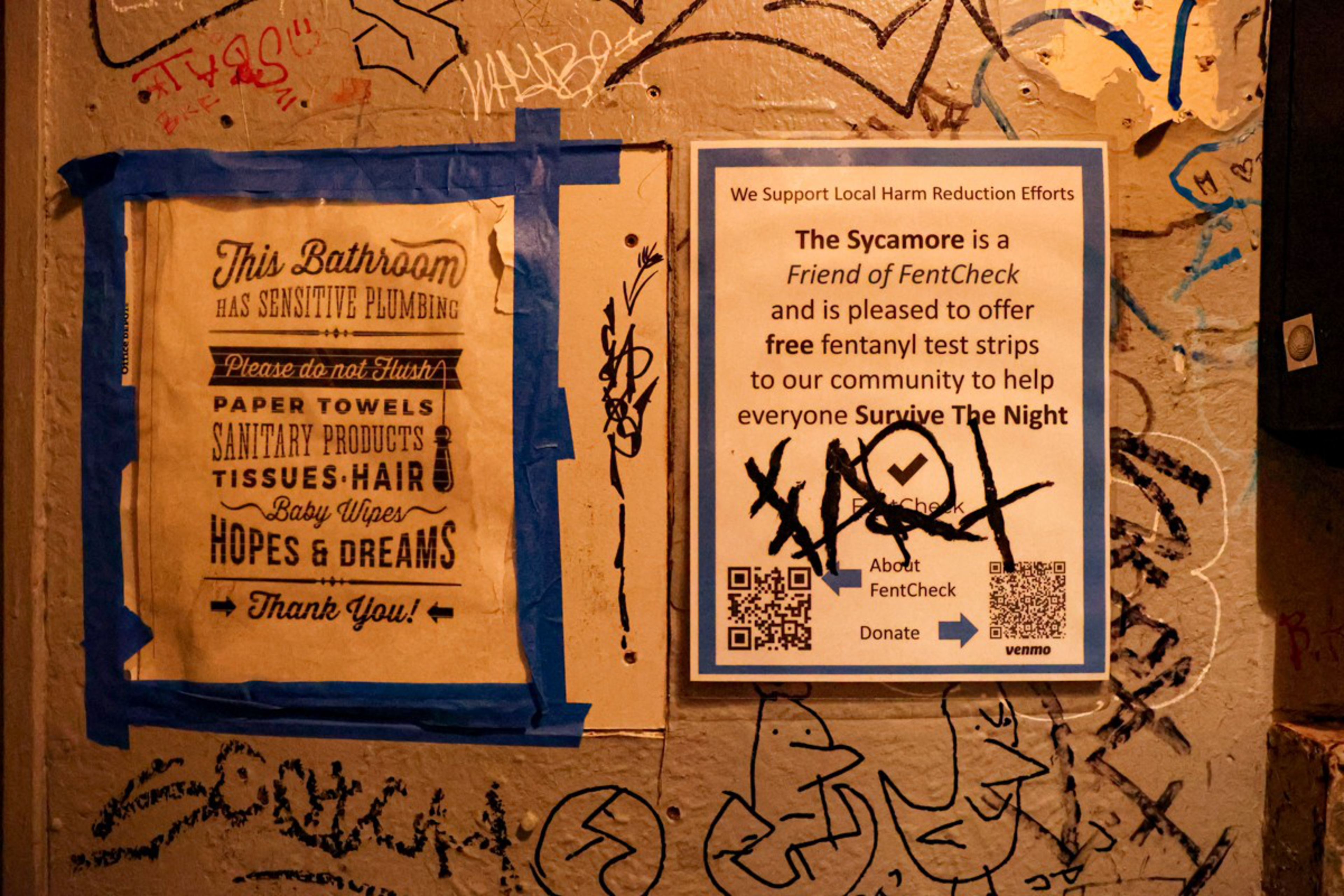 Two signs on a graffiti-covered wall; one asks not to flush items like paper towels and hopes, the other offers free fentanyl test strips for harm reduction.