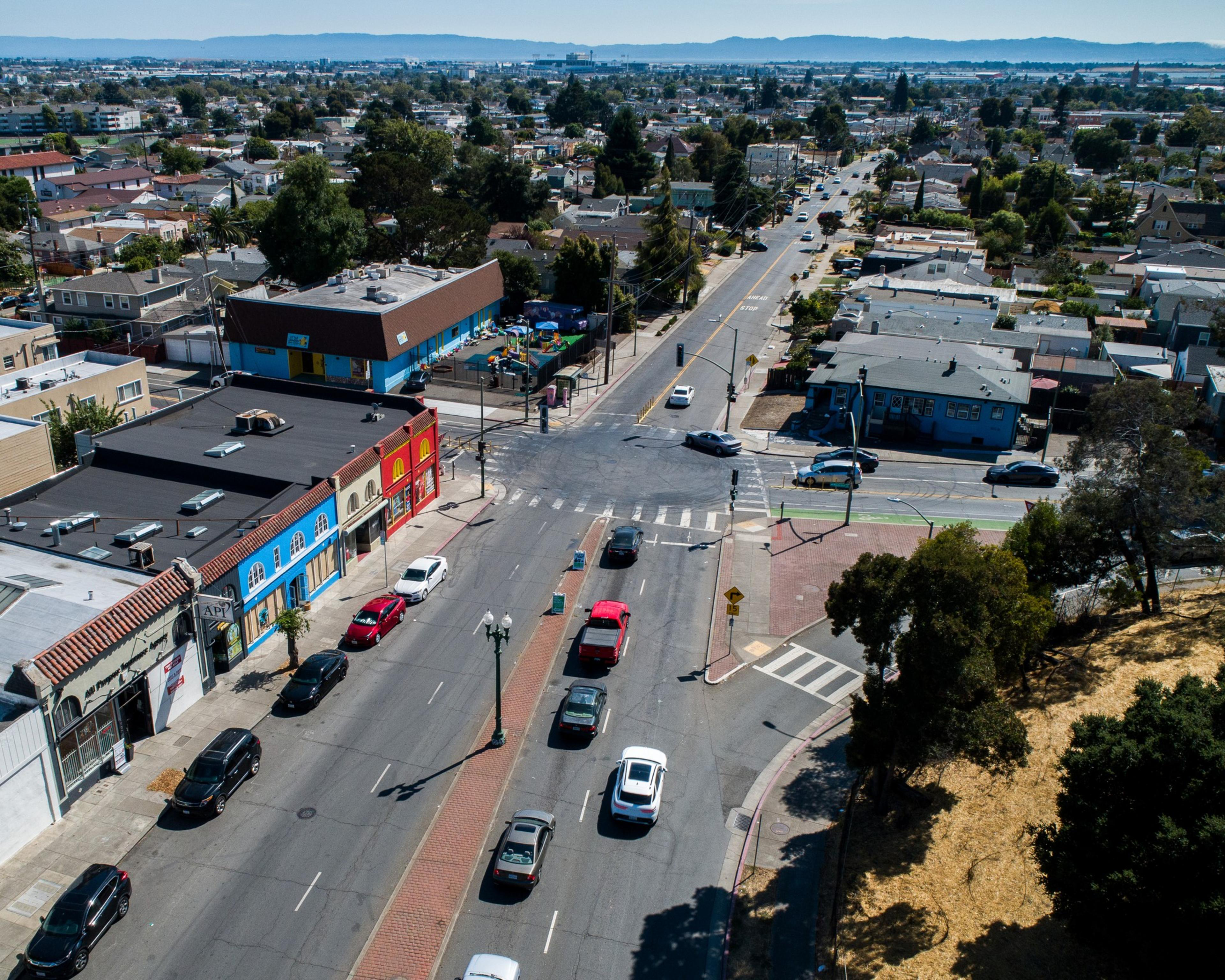 This image depicts a city intersection seen from above, with colorful buildings, parked cars, a playground, houses, and a distant skyline under a clear sky.