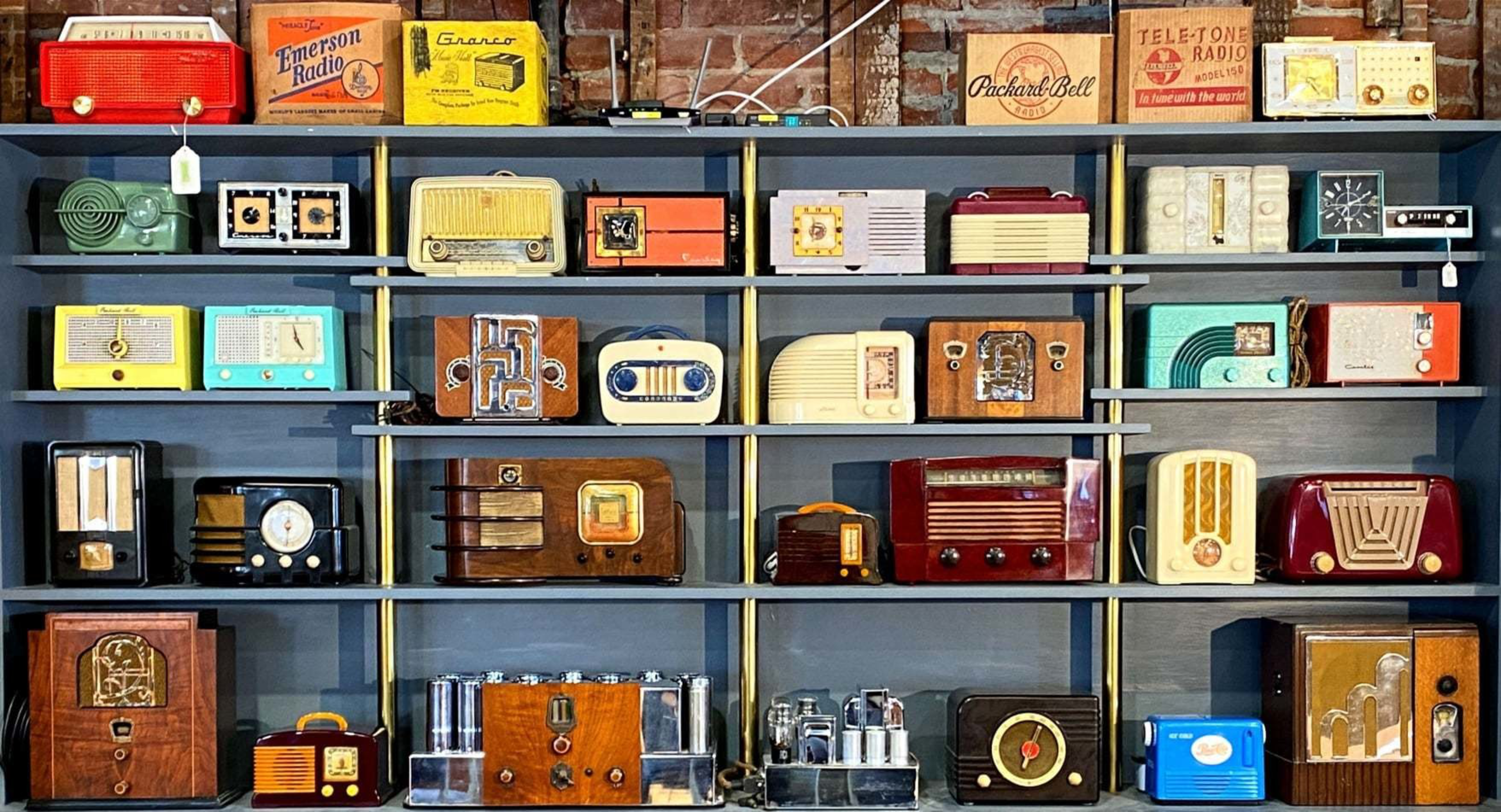 A collection of vintage radios in various colors and styles fills wooden shelves against a brick wall, with some boxed radios on the top shelf.