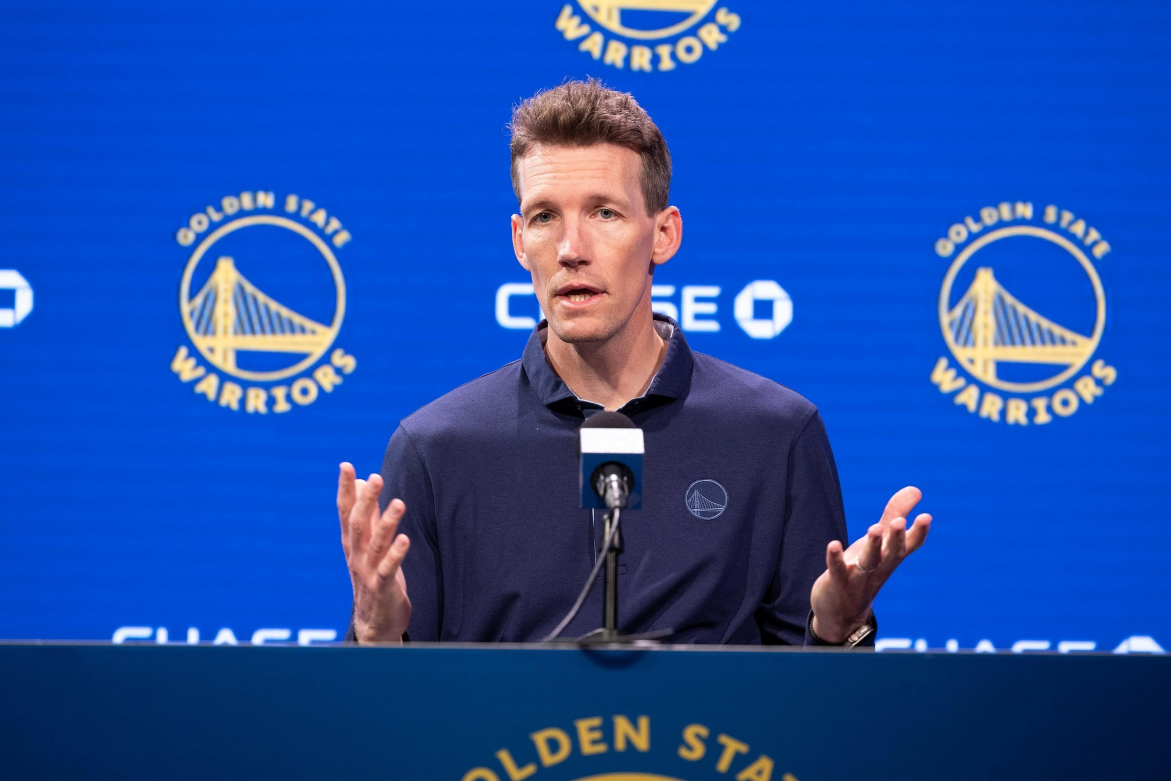 A person stands at a podium with a microphone, speaking. The backdrop features the Golden State Warriors logo repeatedly, and the person wears a navy shirt with the logo.