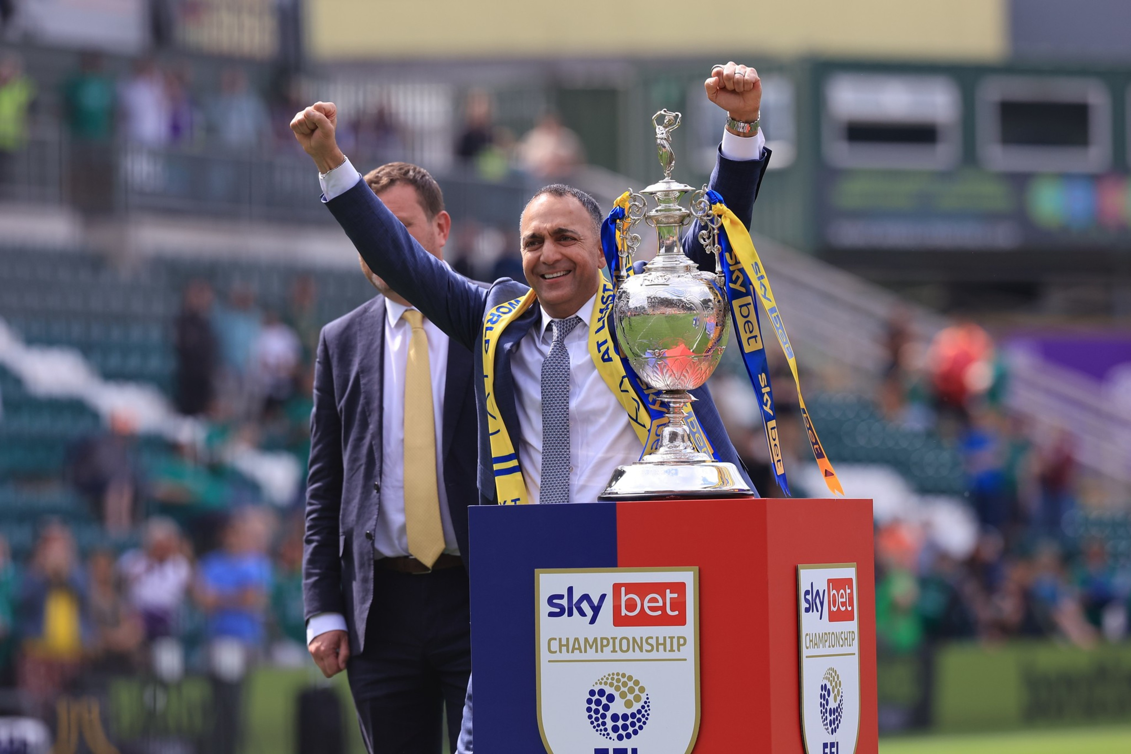 A man in a suit raises both fists in celebration behind a Sky Bet Championship trophy adorned with yellow and blue ribbons.
