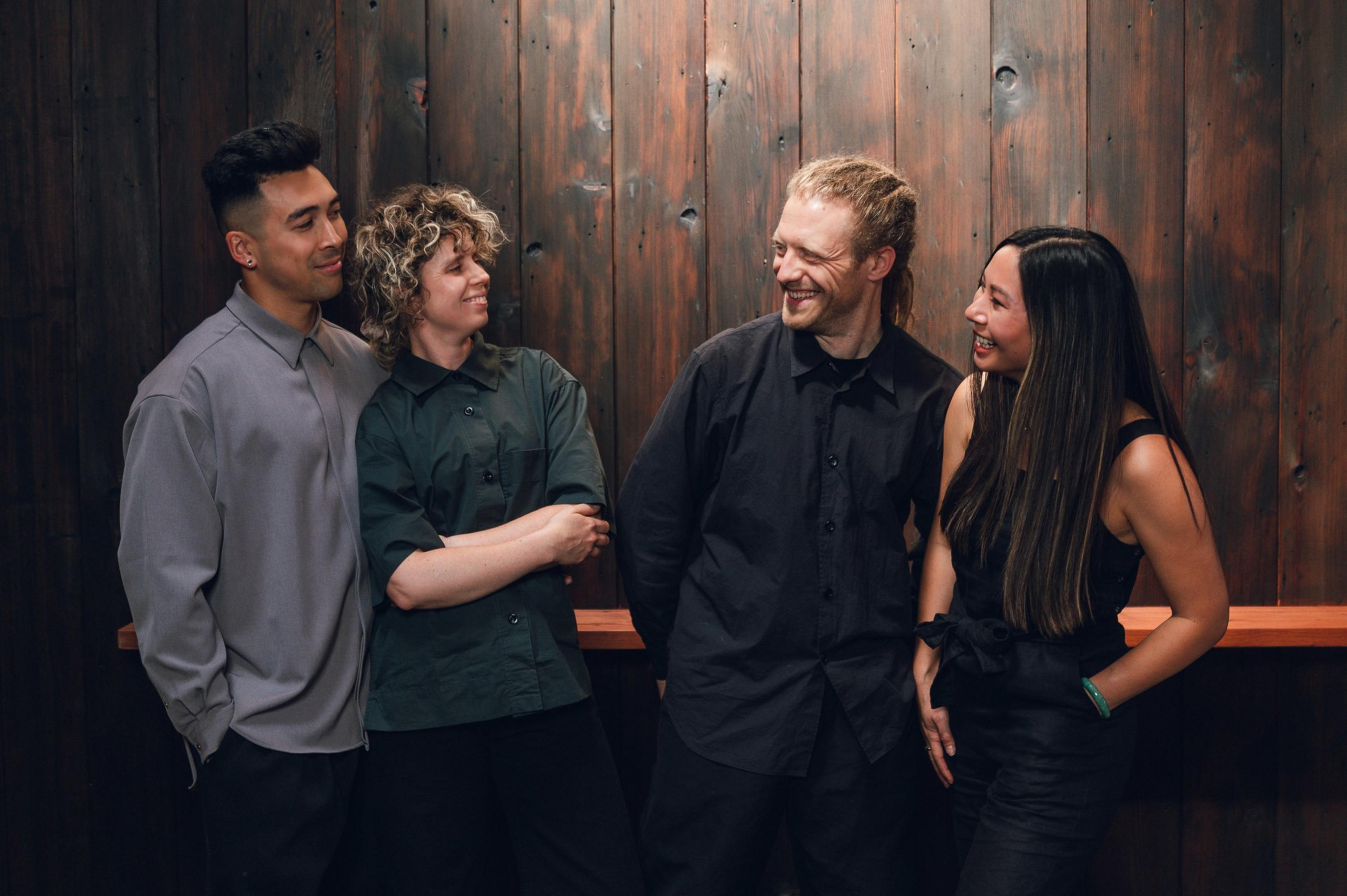 Four friends stand closely together, smiling and engaging warmly against a rustic wooden wall background.