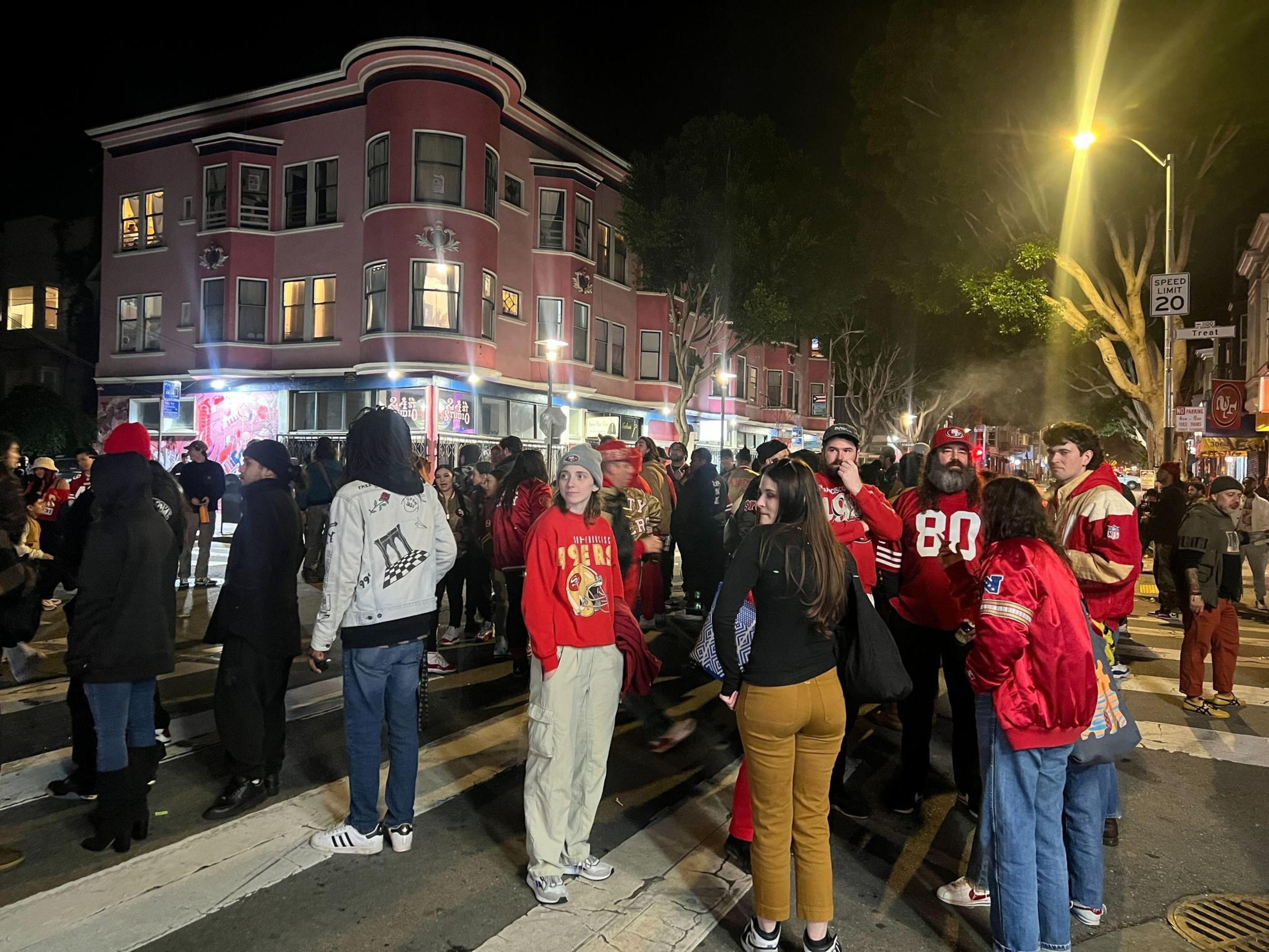A bustling crowd of people, many in red attire, gathers at a night-time street intersection with a pink building backdrop.