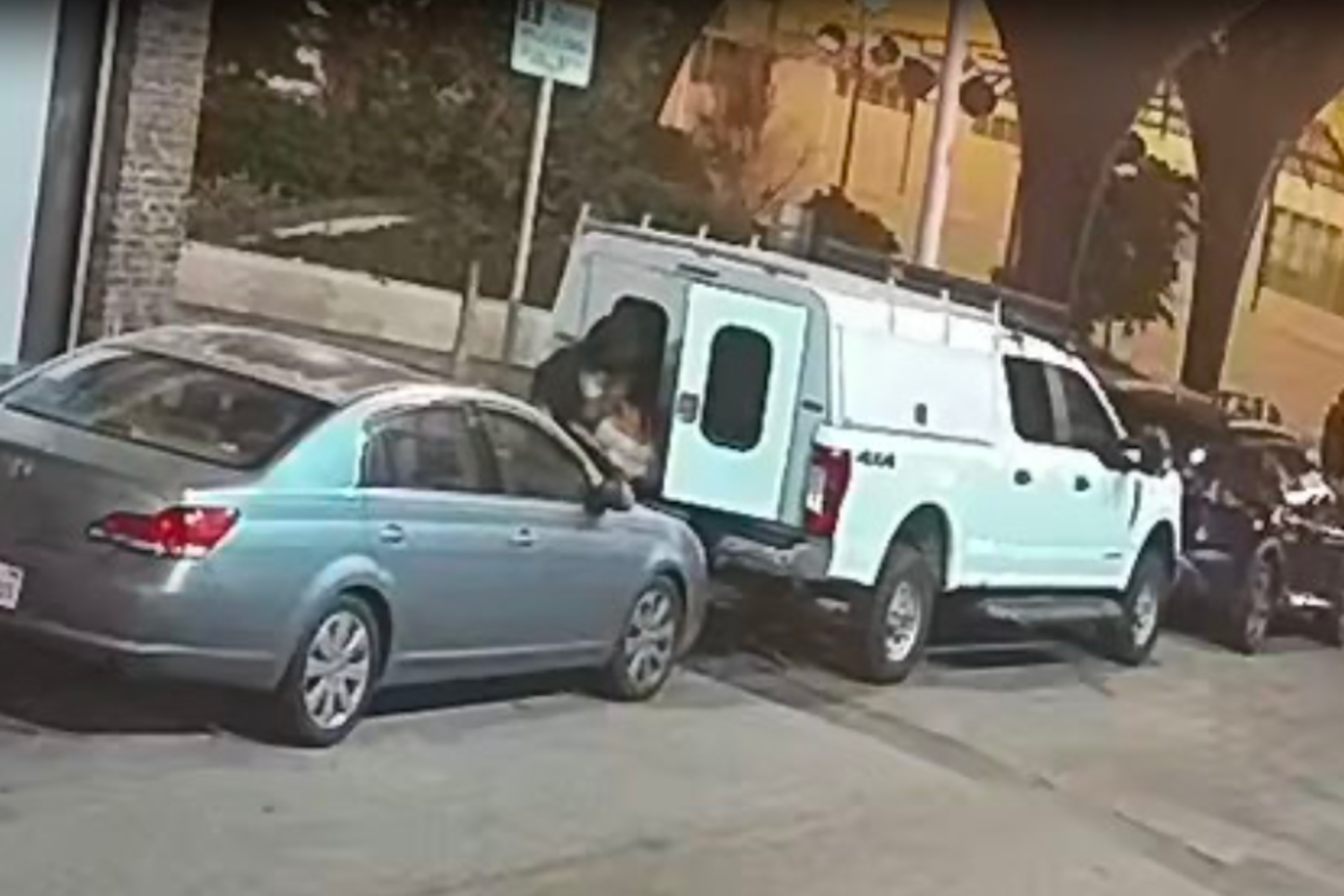 A person appears to be breaking into a parked white truck at night in a residential area, with other cars and a couple of people visible in the background.