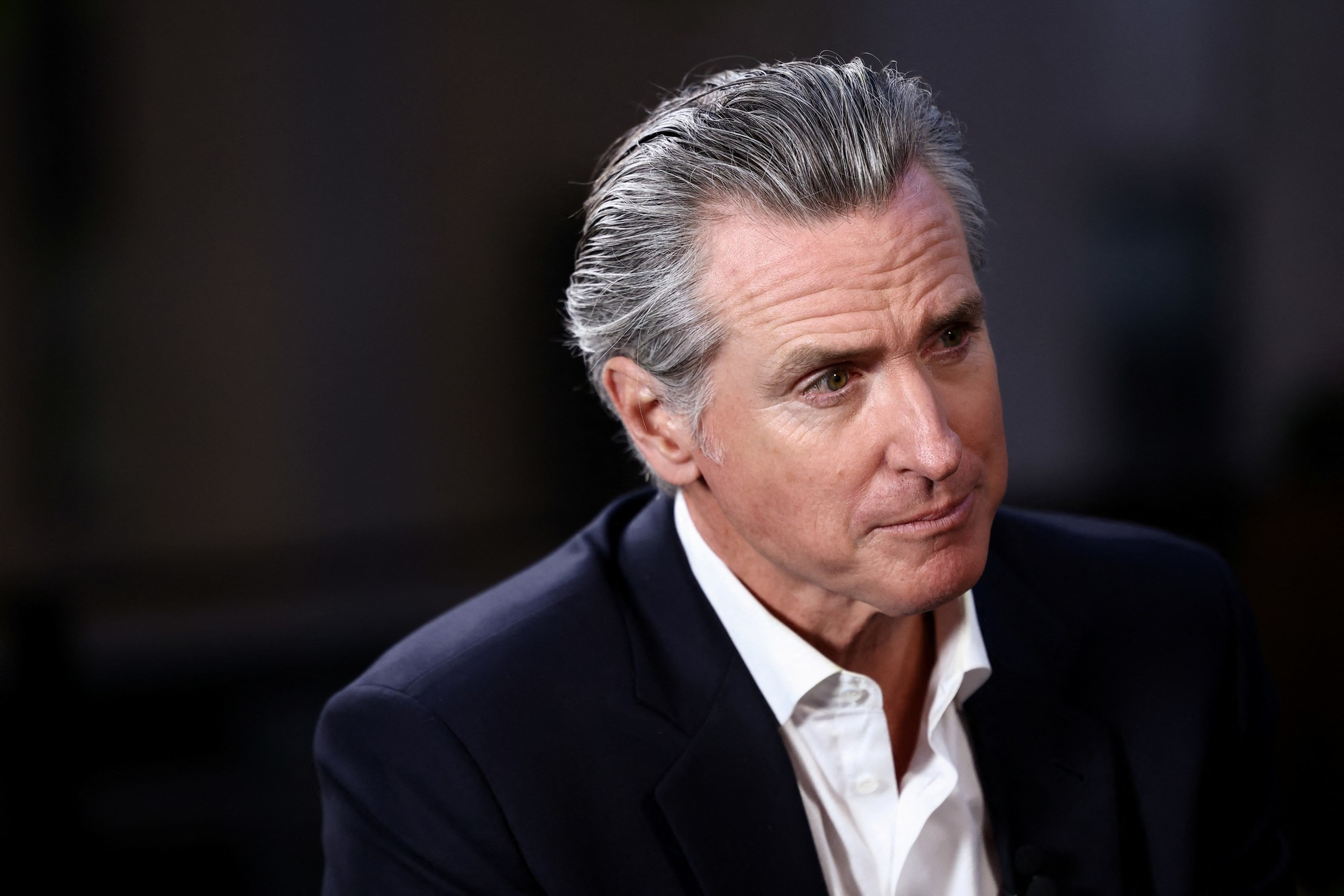 A middle-aged man with slicked-back gray hair wears a dark suit jacket and a white shirt, looking thoughtfully to his right.