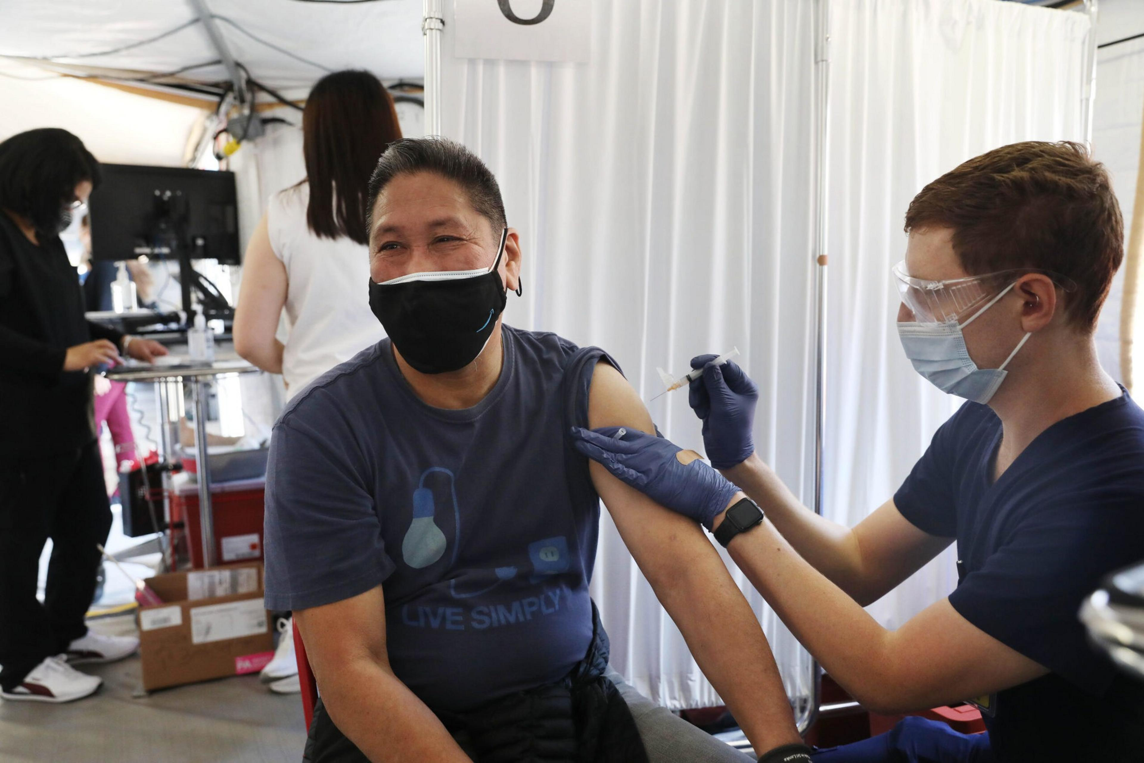 A person wearing a mask is smiling while receiving a vaccine shot in their upper arm from a masked healthcare worker wearing gloves and protective eyewear.