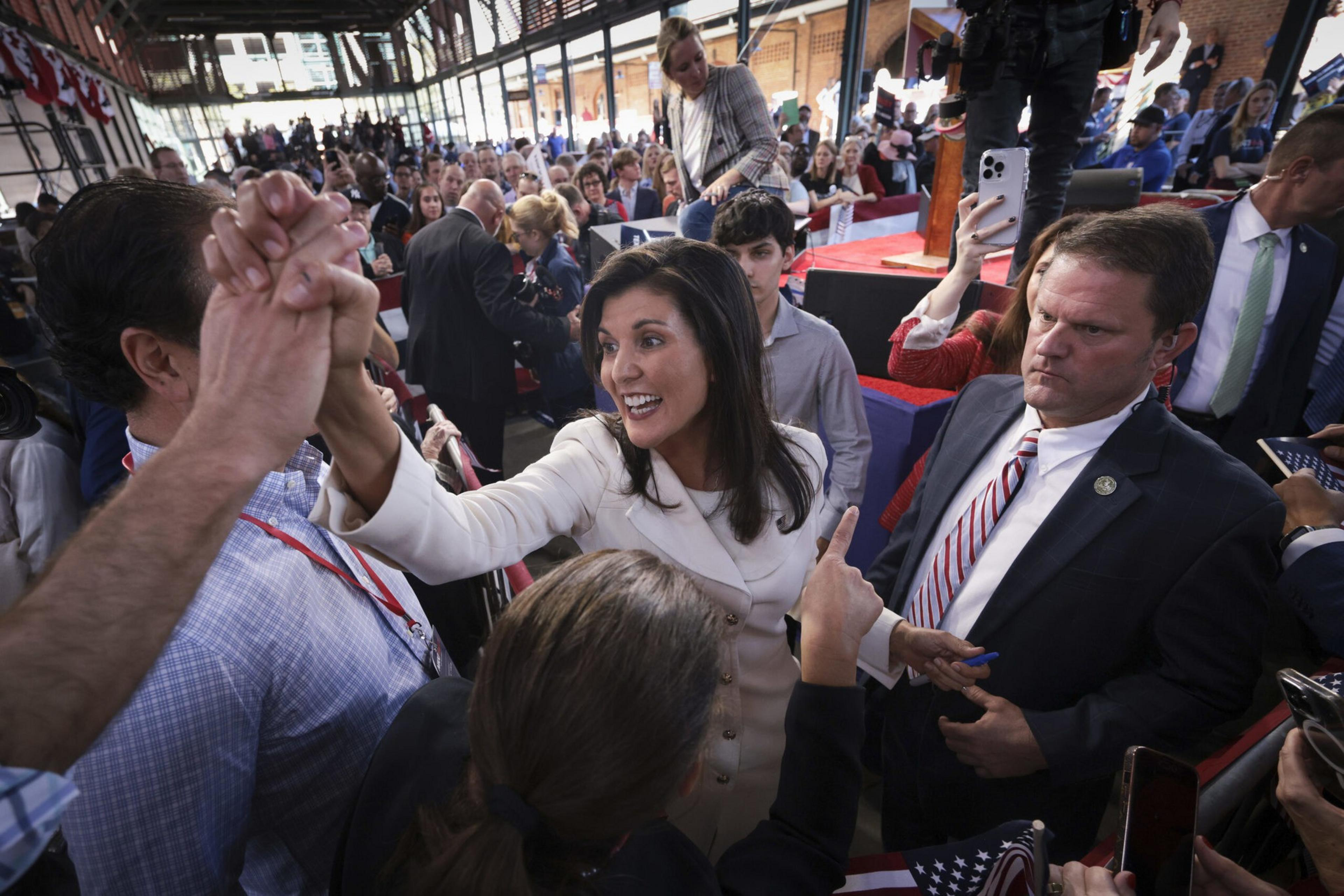 A woman in a white coat energetically shakes hands with a crowd, while a serious man in a suit stands beside her and people hold up phones to record.