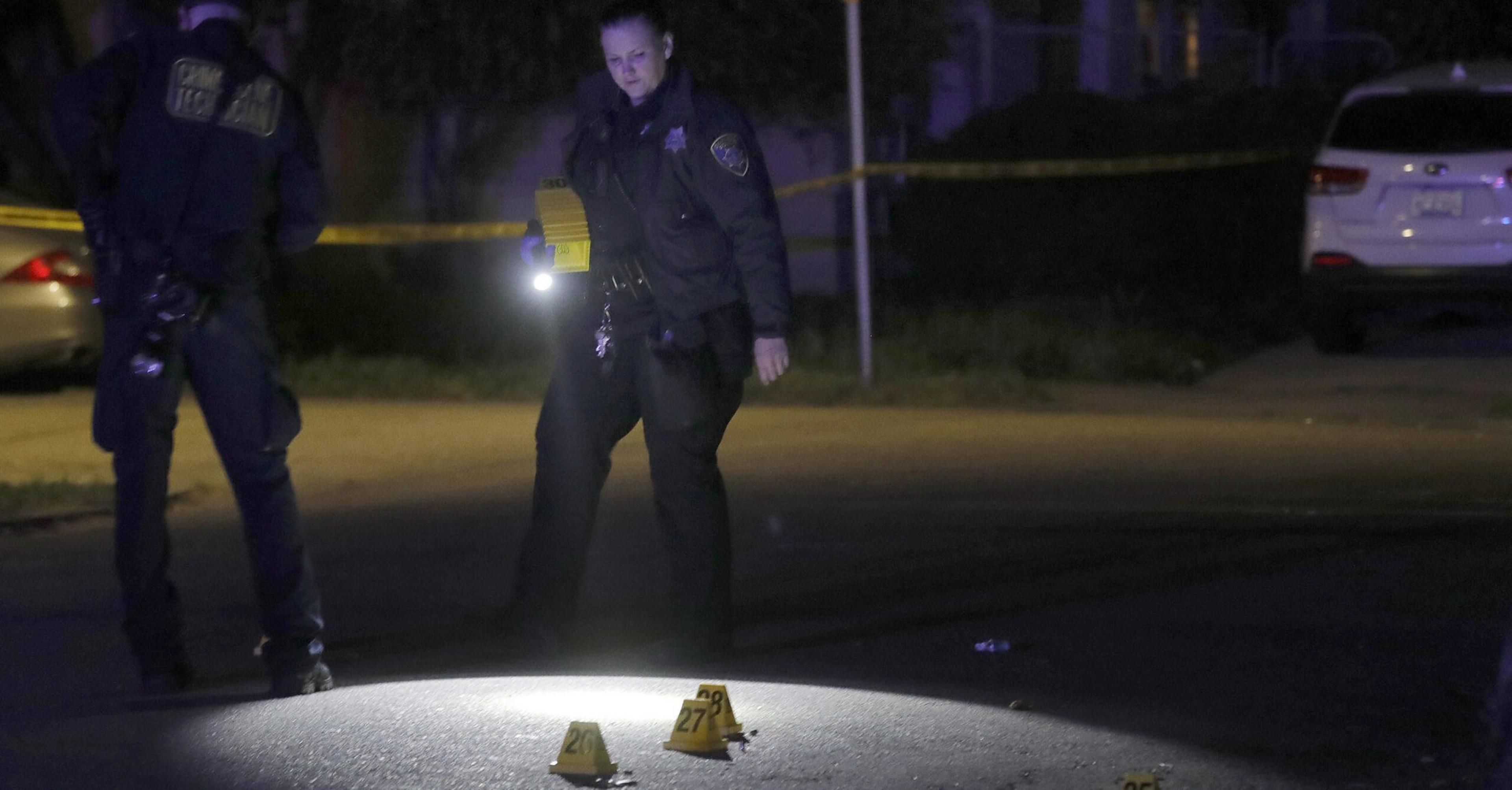 A police officer shines a flashlight on numbered evidence markers on a street at night, with another officer and police tape visible in the background.