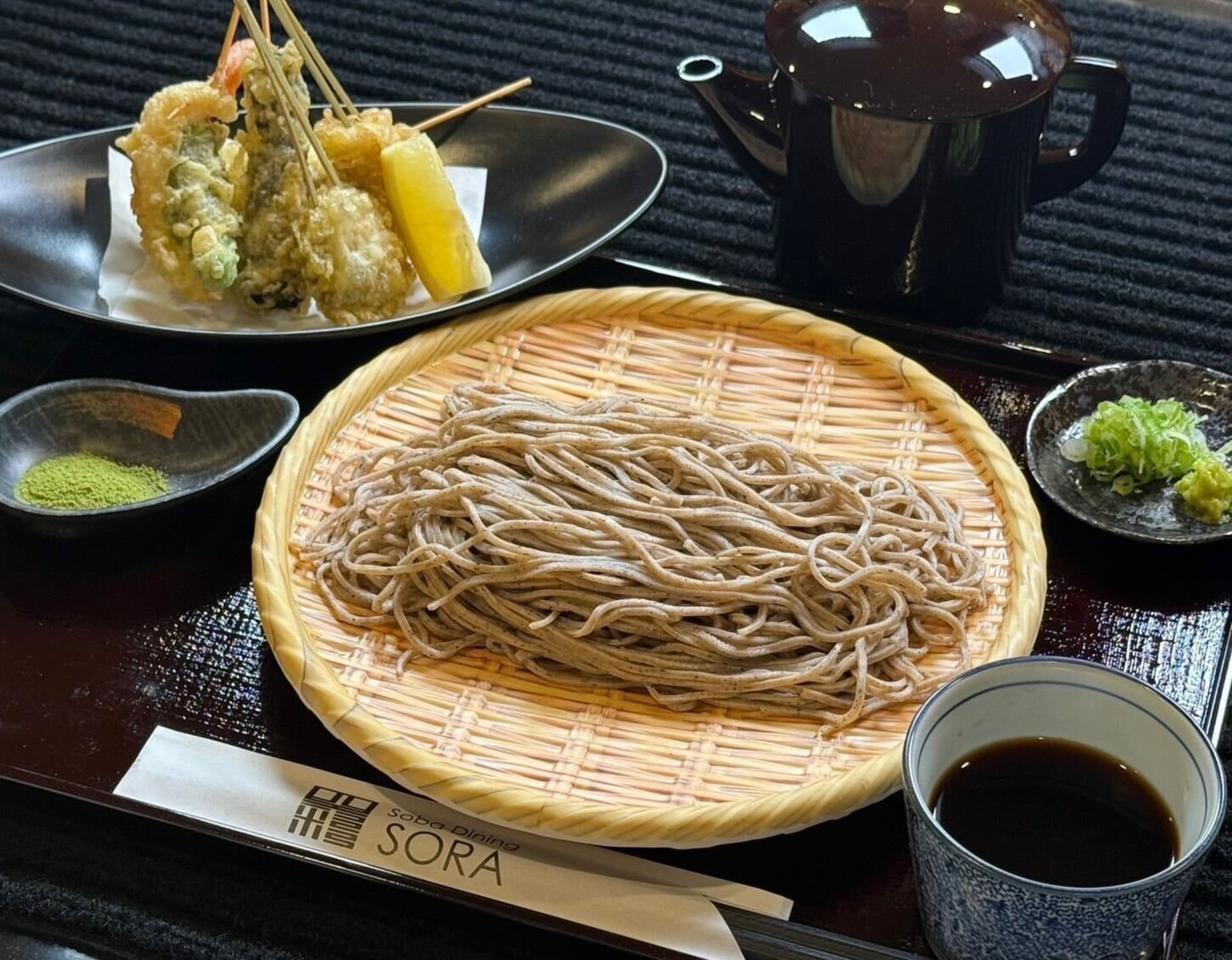 Cold soba noodles on a bamboo tray, served with tempura skewers, green tea powder, dipping sauce, and garnishes of green onion and wasabi.