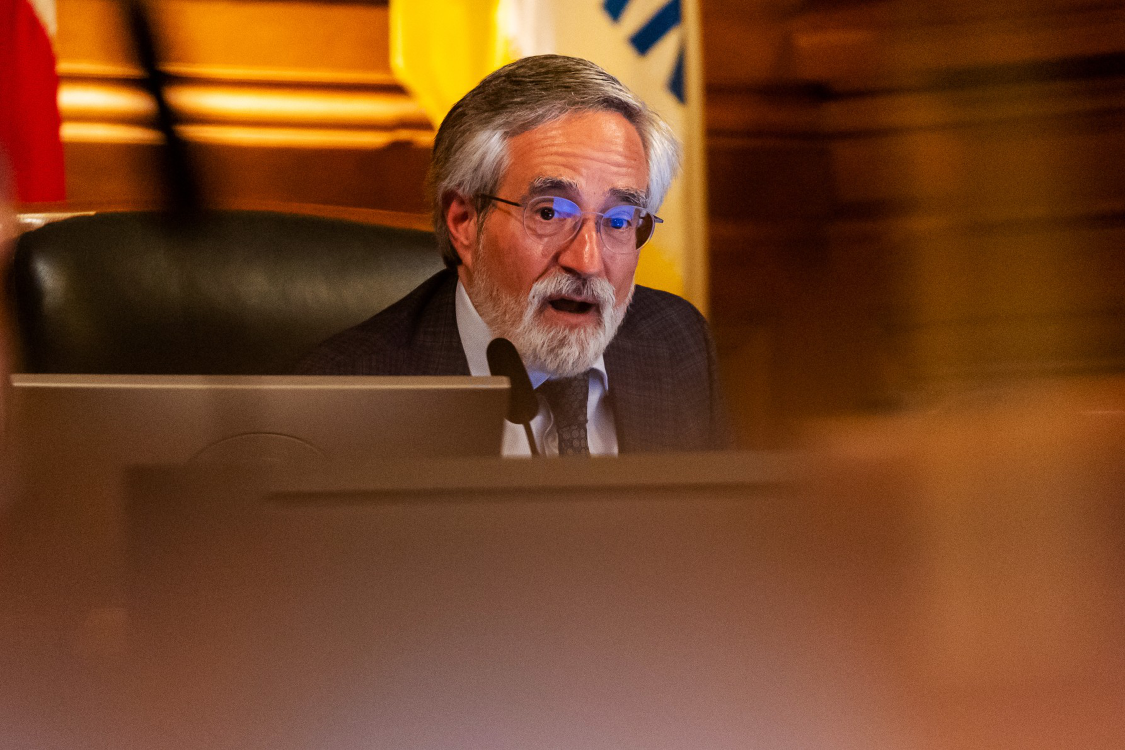 An older man with gray hair and beard, wearing glasses and a suit, speaks into a microphone while seated behind a desk with a computer monitor.