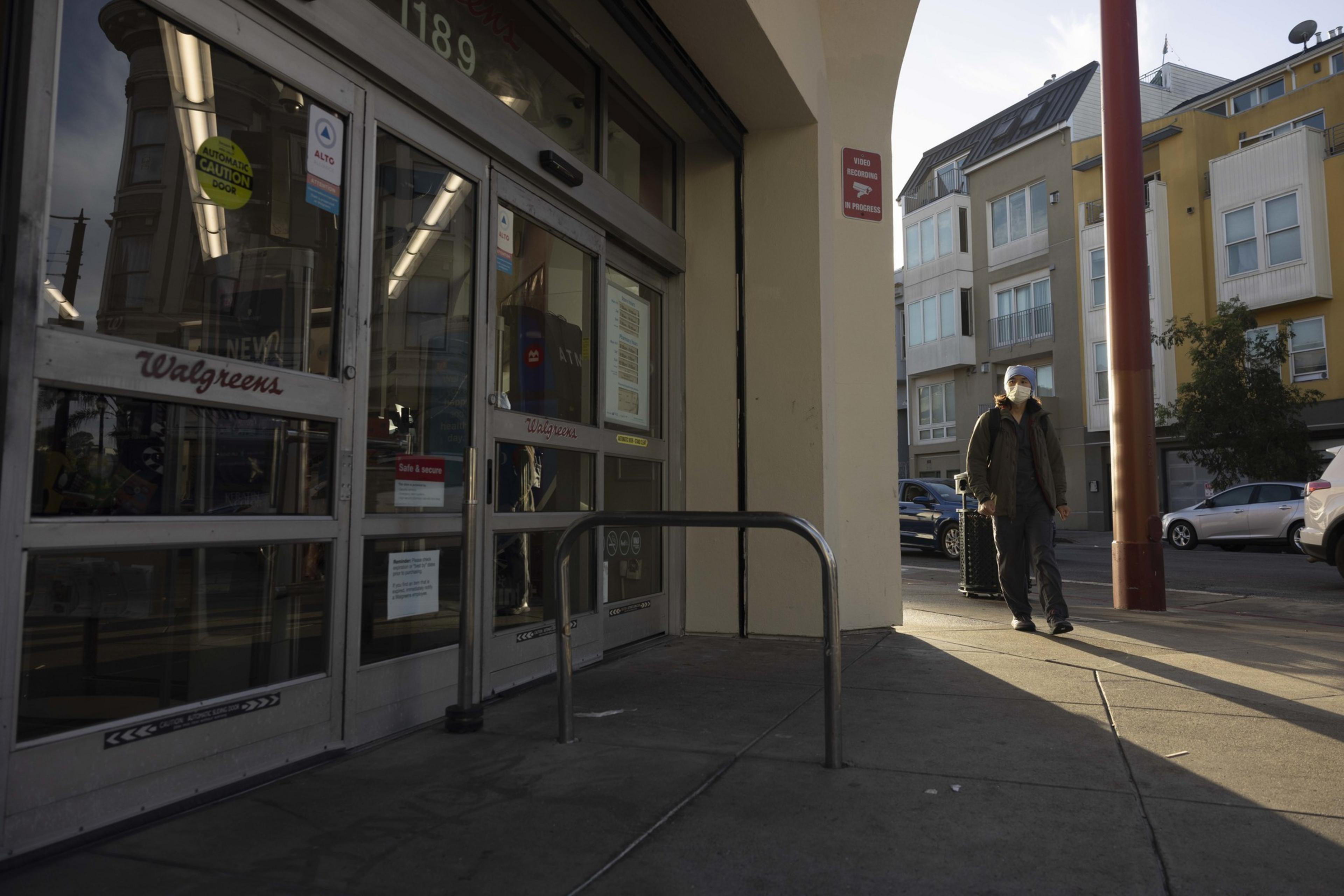 A person wearing a mask walks past a Walgreens store entrance on a city street with buildings and parked cars nearby. The lighting suggests late afternoon.