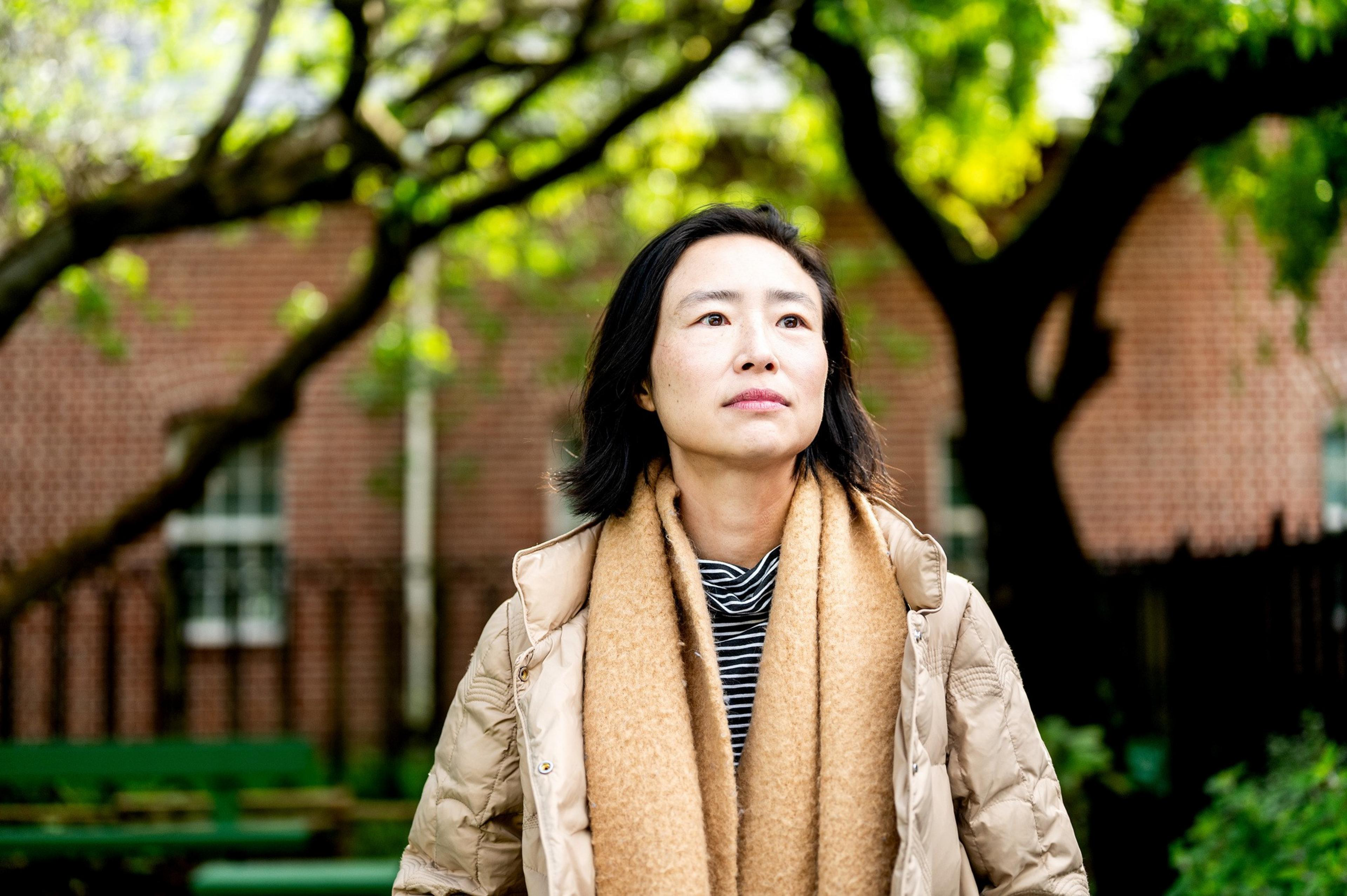 A person stands outside, wearing a tan jacket and scarf, with a thoughtful expression. Trees and a brick wall form the background, creating a serene setting.