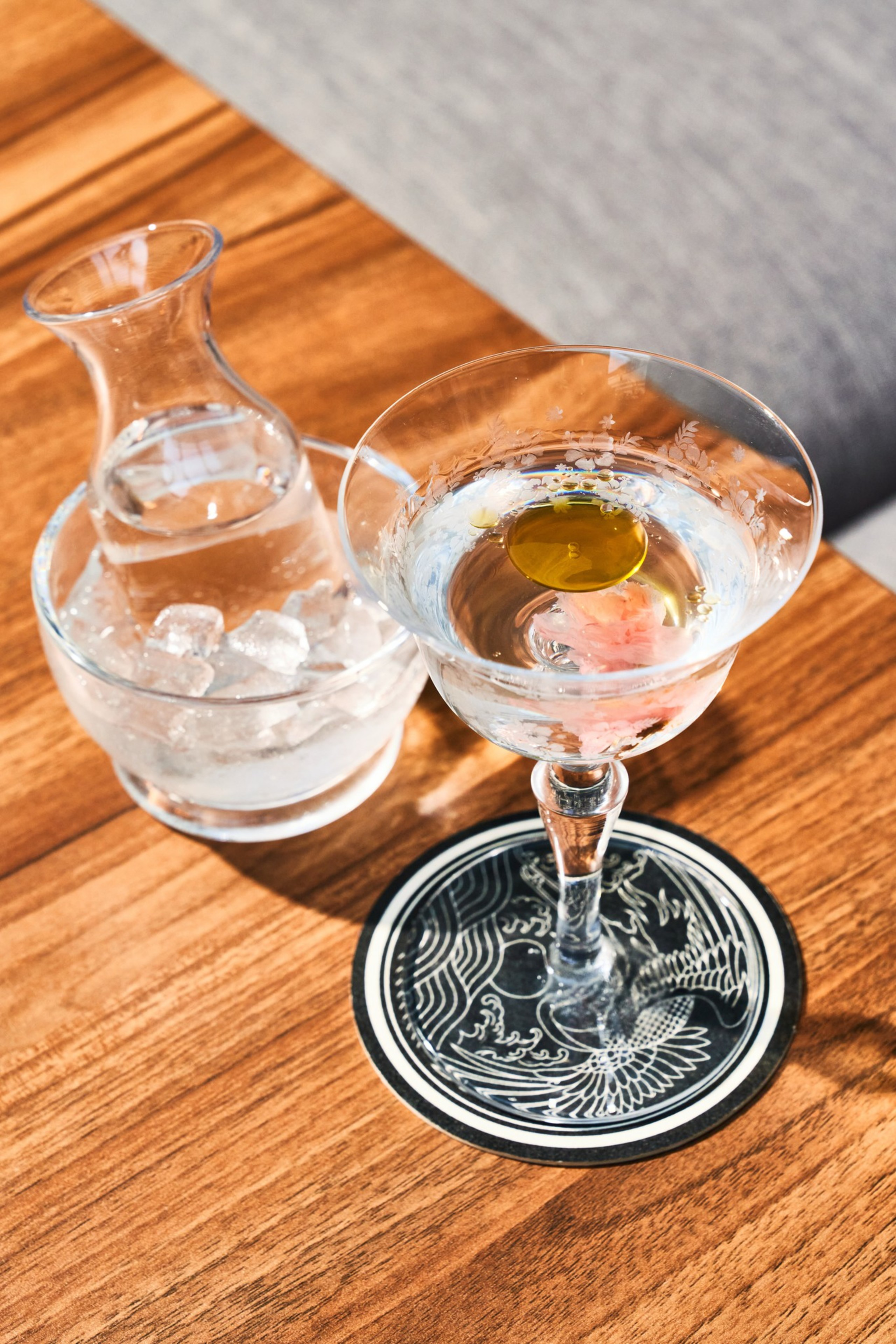 A martini glass with a clear liquid and a yellow olive in it sits on a patterned coaster. Nearby, a small carafe is placed in a bowl of ice on a wooden table.