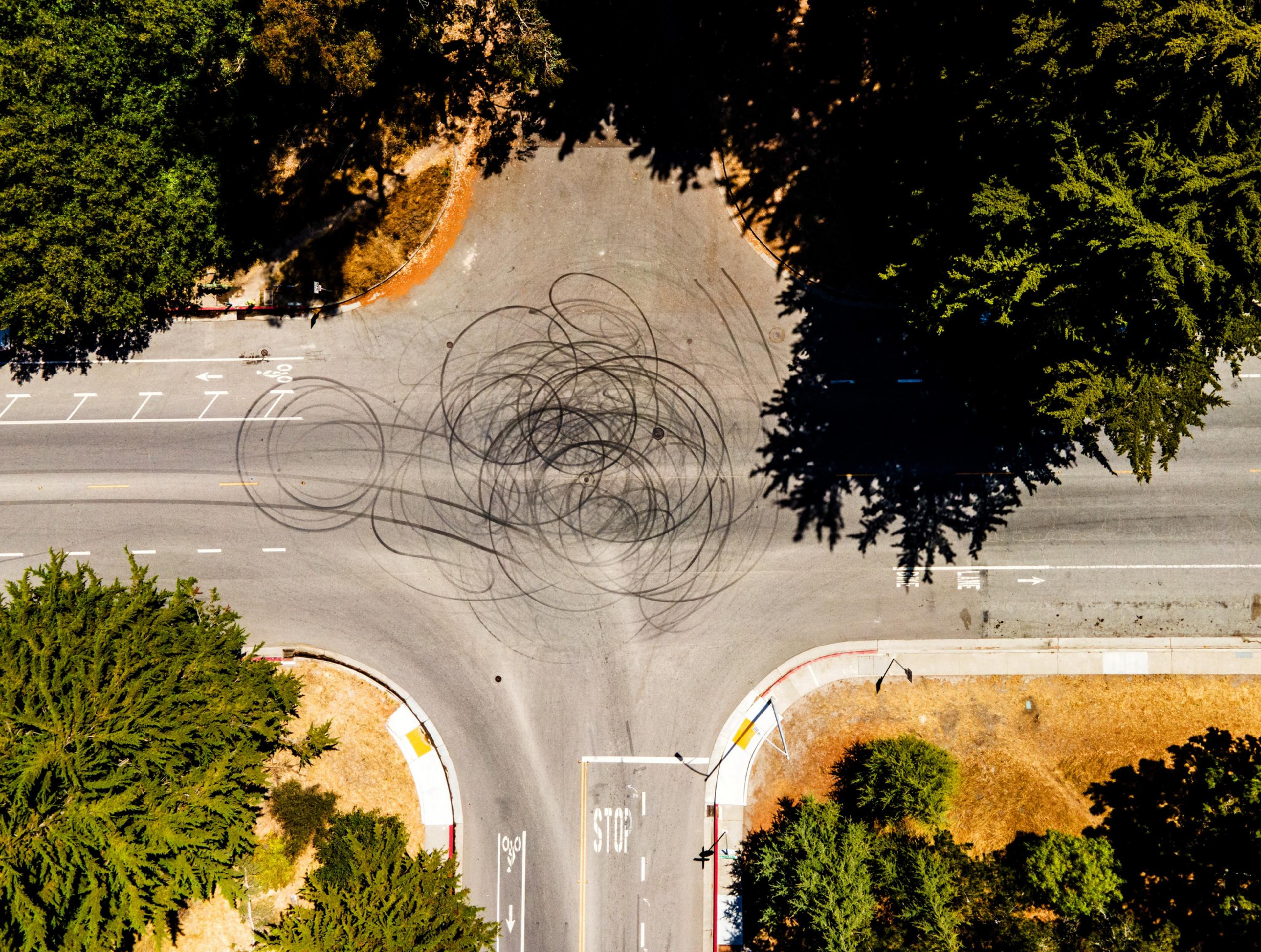 An aerial view of an intersection shows dark tire skid marks in circular patterns on the road. The area is surrounded by trees and some dry grass patches.