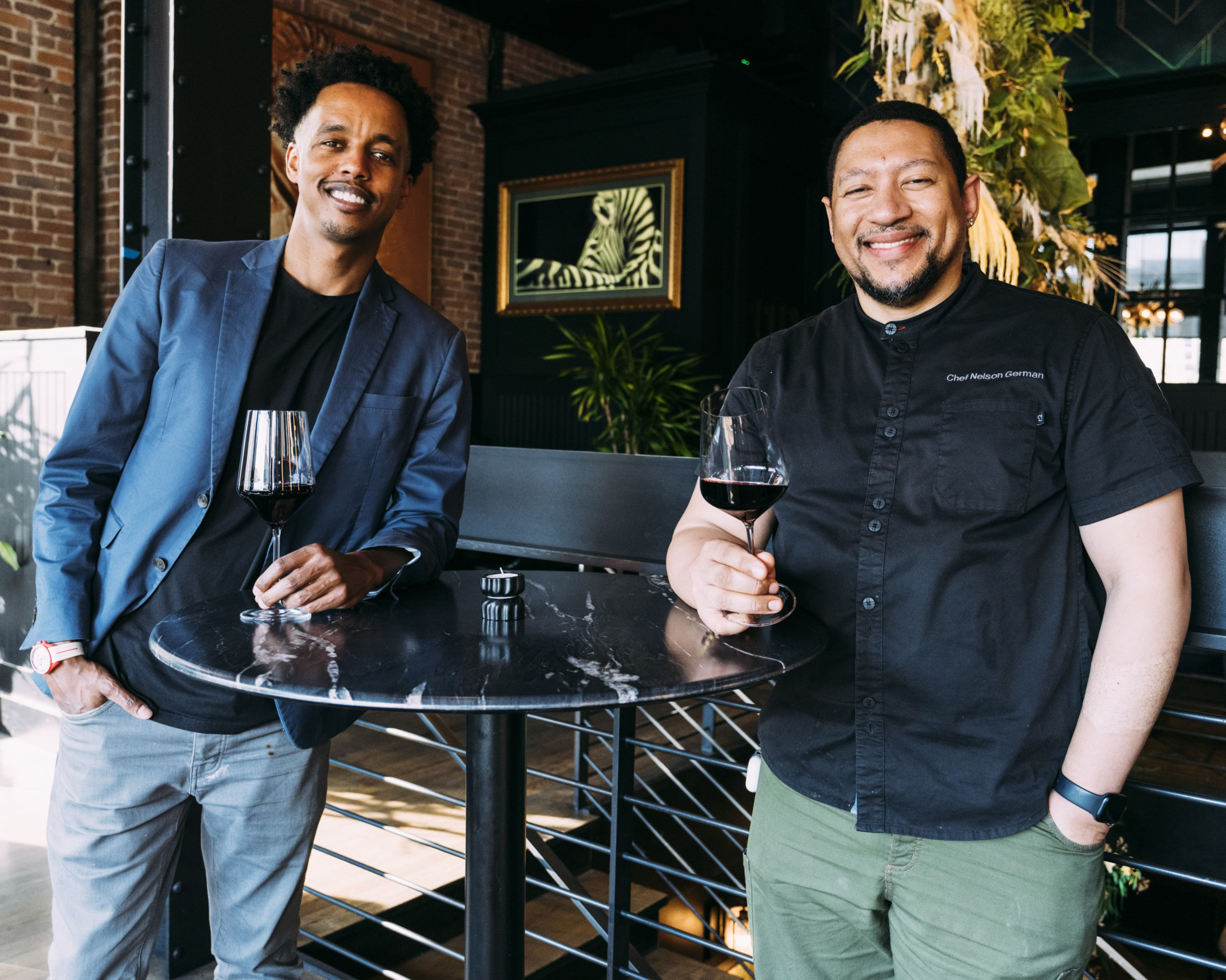Two men are smiling while leaning on a black marble table, each holding a glass of red wine. One wears a blue blazer, and the other is in a black chef's shirt.