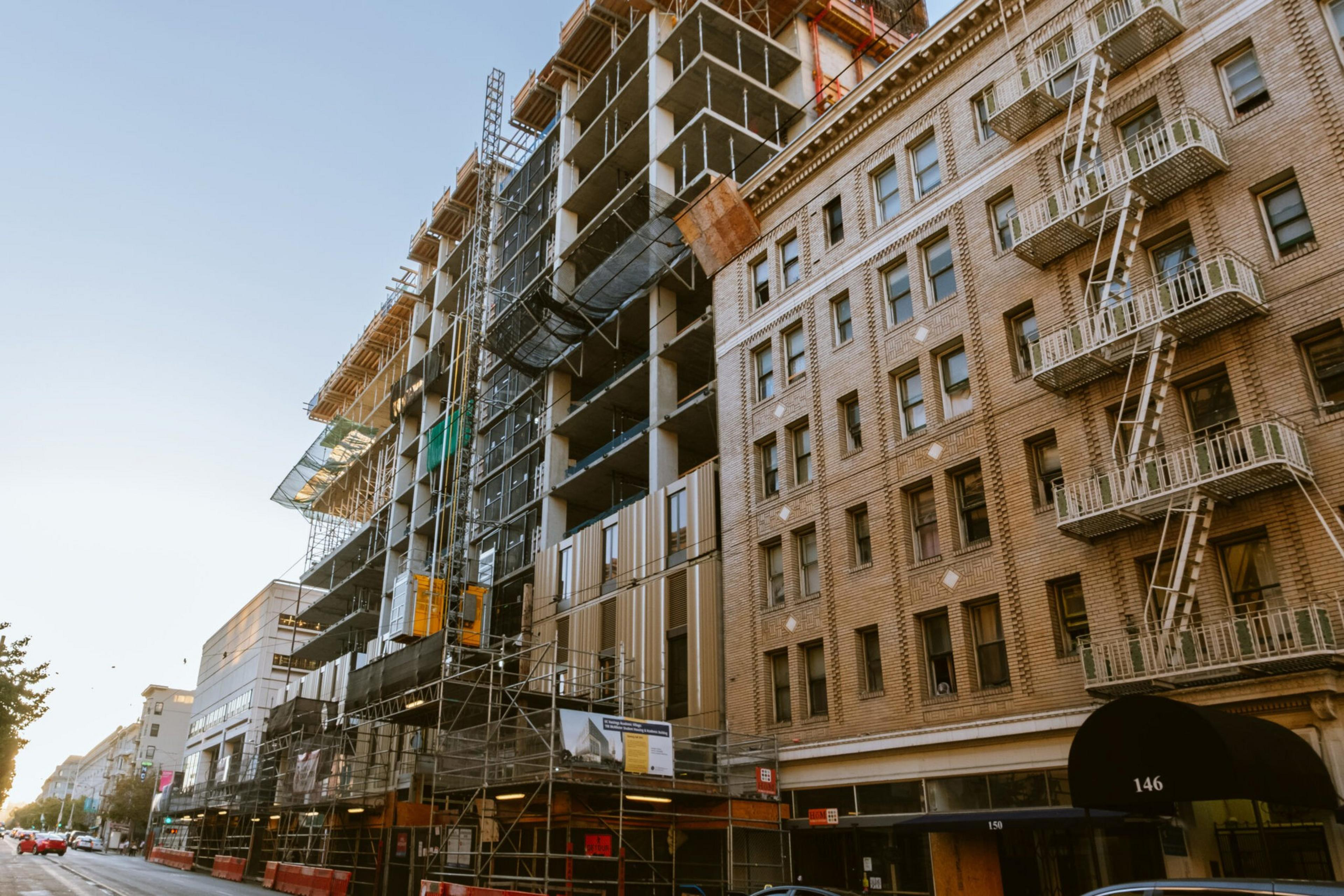 A multi-story building under construction stands next to an older brick building with fire escapes, lining a city street at sunset.