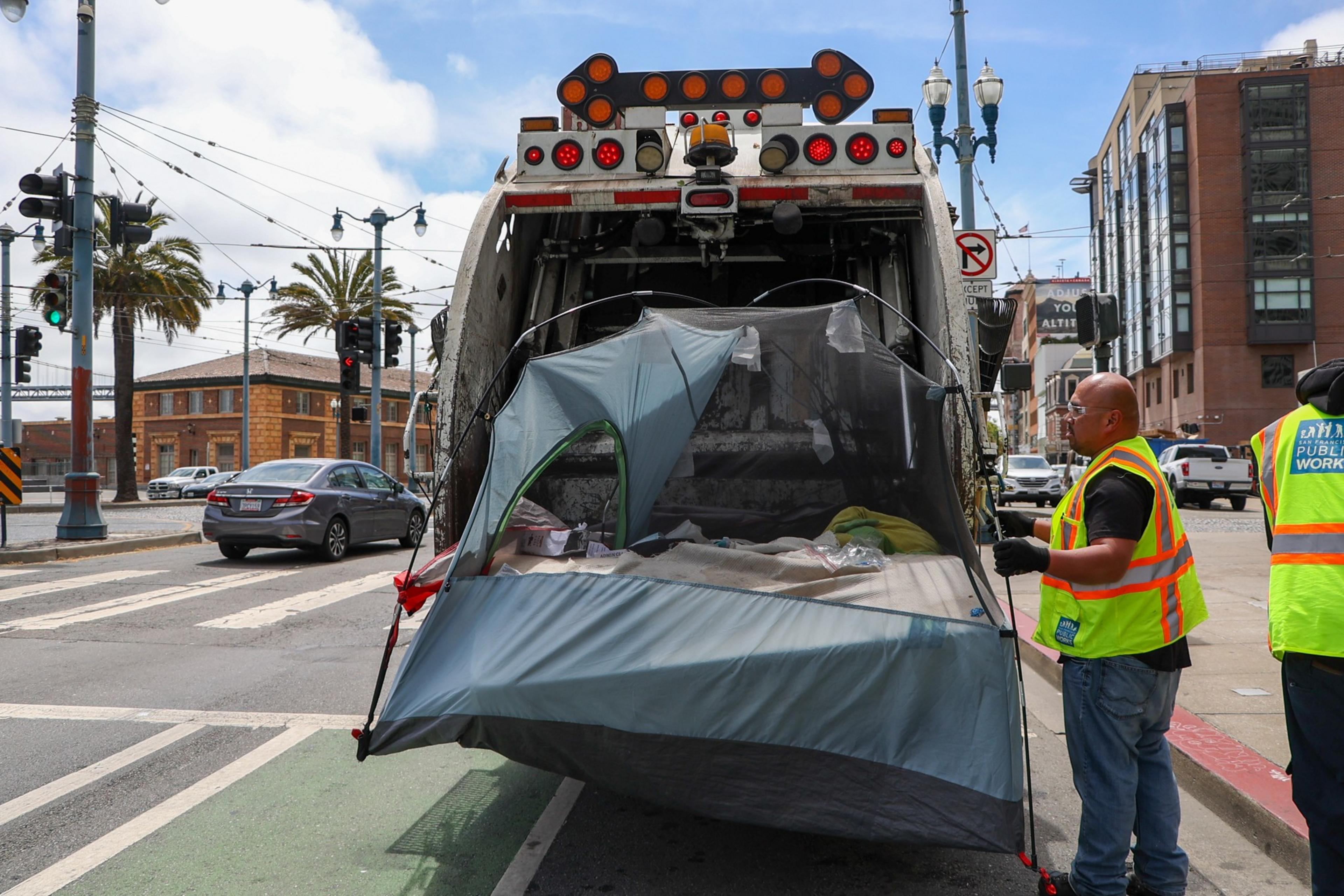 A person in a safety vest dismantles a tent stuck in the back of a garbage truck on a city street with buildings and cars nearby.