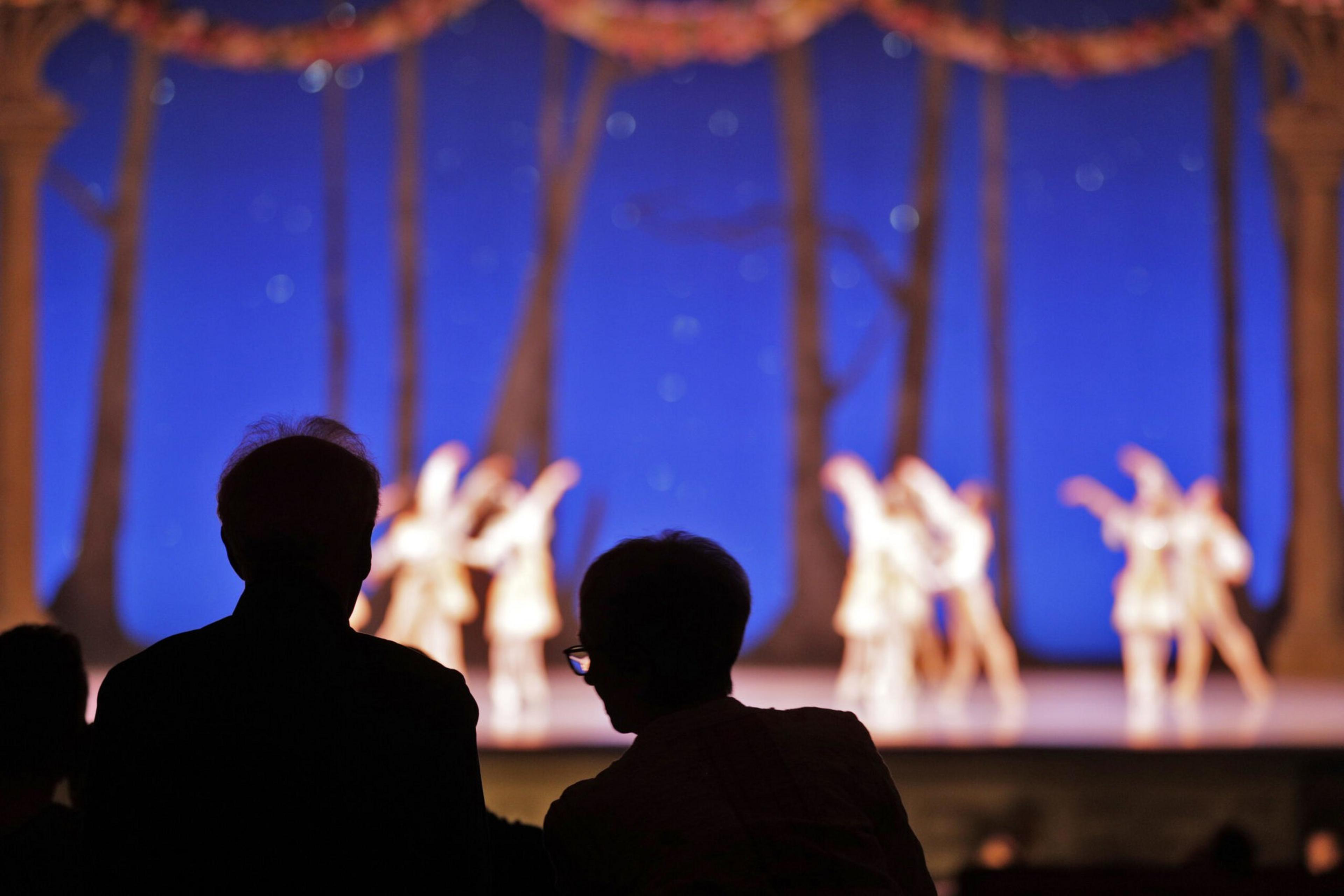 Two people sit in silhouette watching a ballet performance on stage with dancers dressed in white against a blue backdrop with trees.