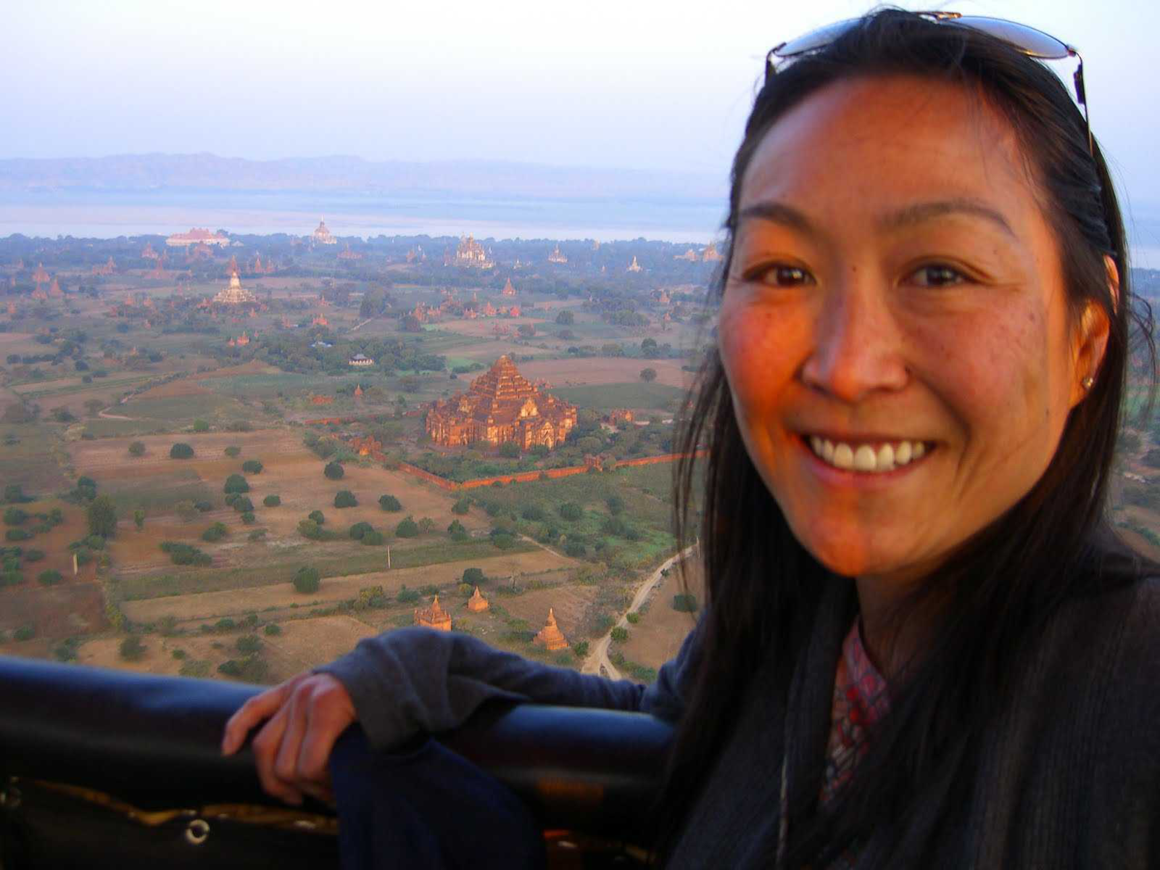 A smiling woman with sunglasses on her head leans on a railing overlooking a vast landscape dotted with ancient temples and greenery.
