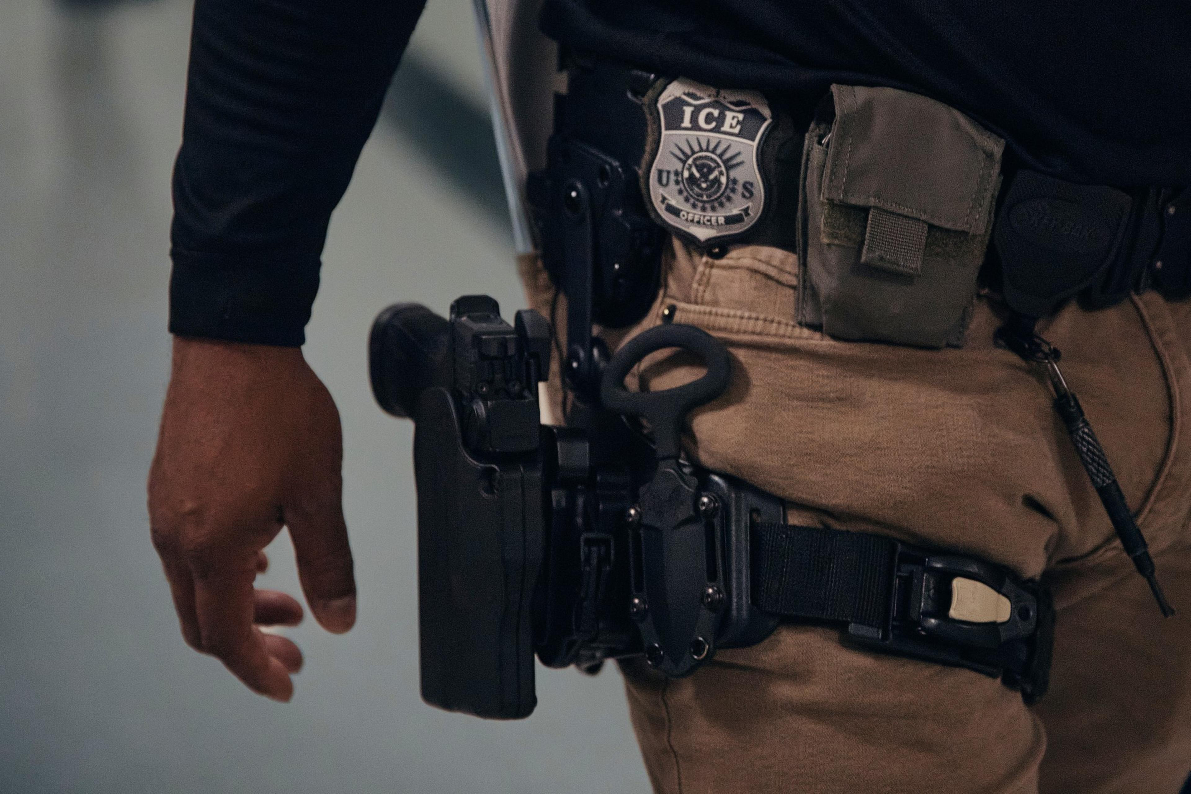 A close-up of a person's waist shows a badge labeled "ICE" on a belt, surrounded by tactical gear including a holstered gun, a pouch, and tools.