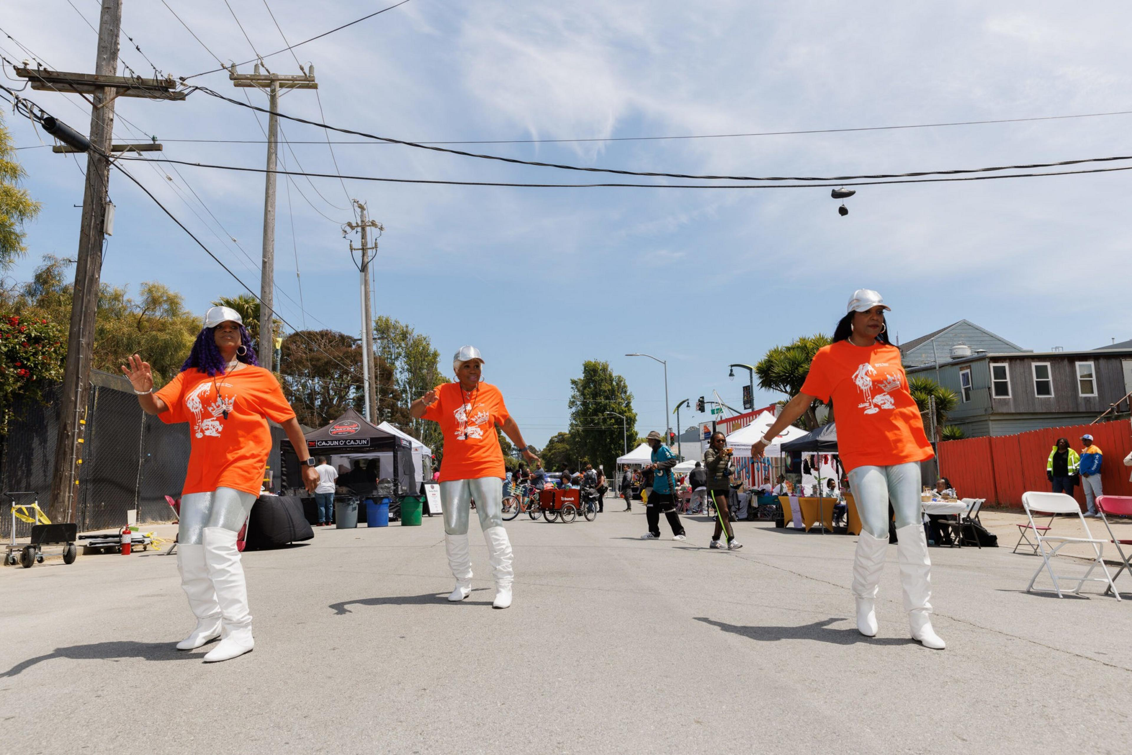 Three people dancing on a street in matching orange shirts and white pants with bystanders and booths in the background.