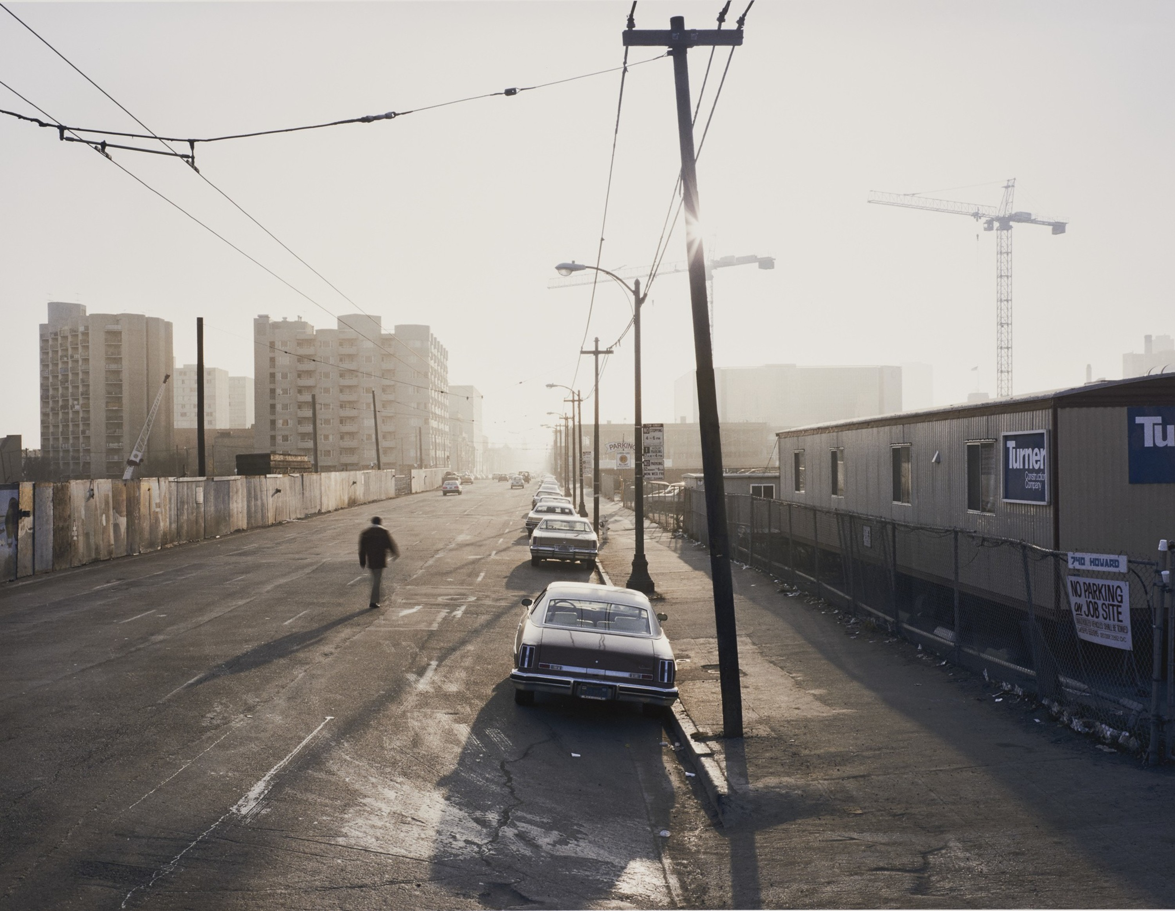 A nearly empty street lined with parked cars, a lone pedestrian walking, utility poles casting long shadows, and buildings under a hazy, bright sky.