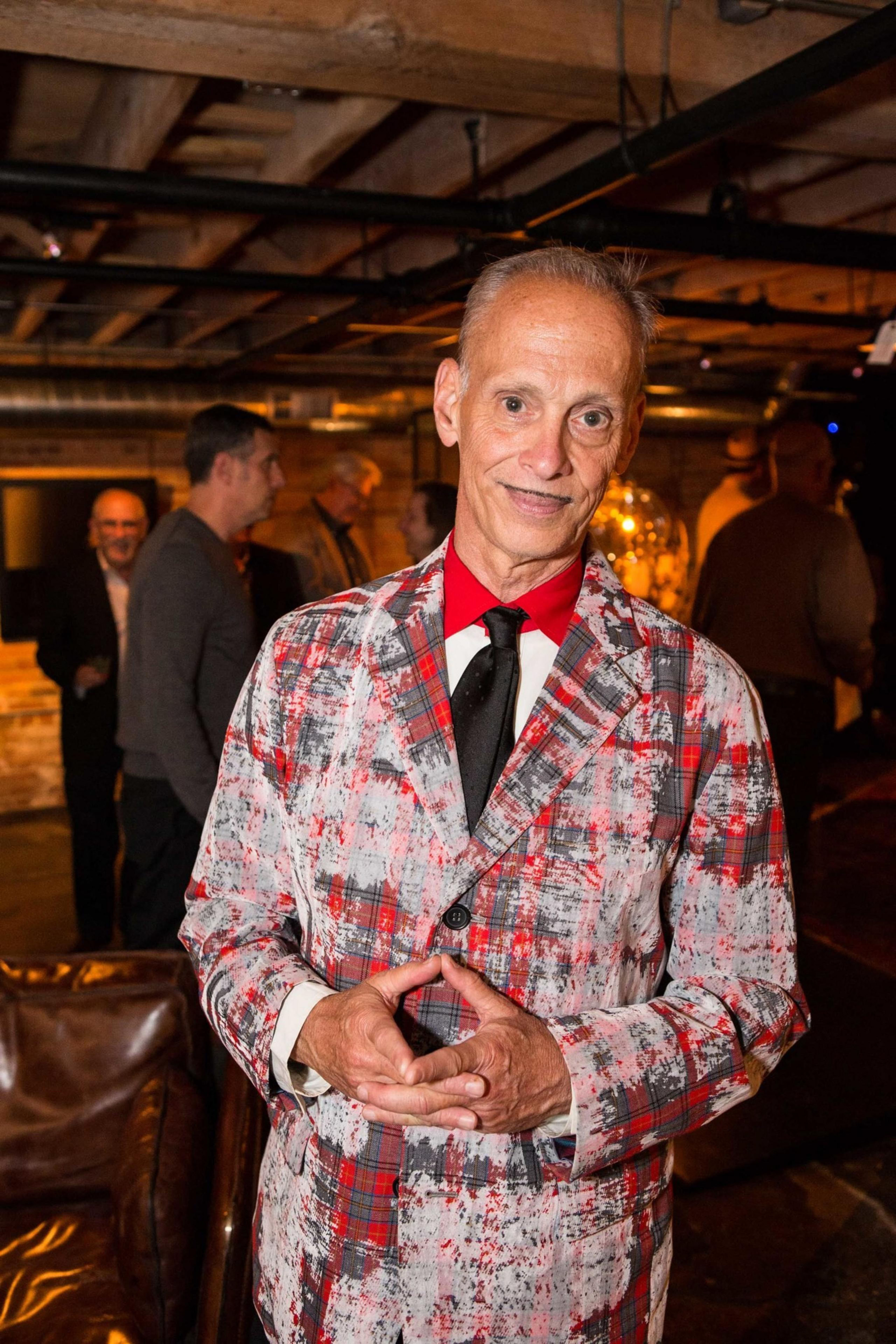 A man in a colorful, abstract-patterned suit and red shirt stands indoors, smiling with hands clasped. Several people are conversing in the dimly-lit background.