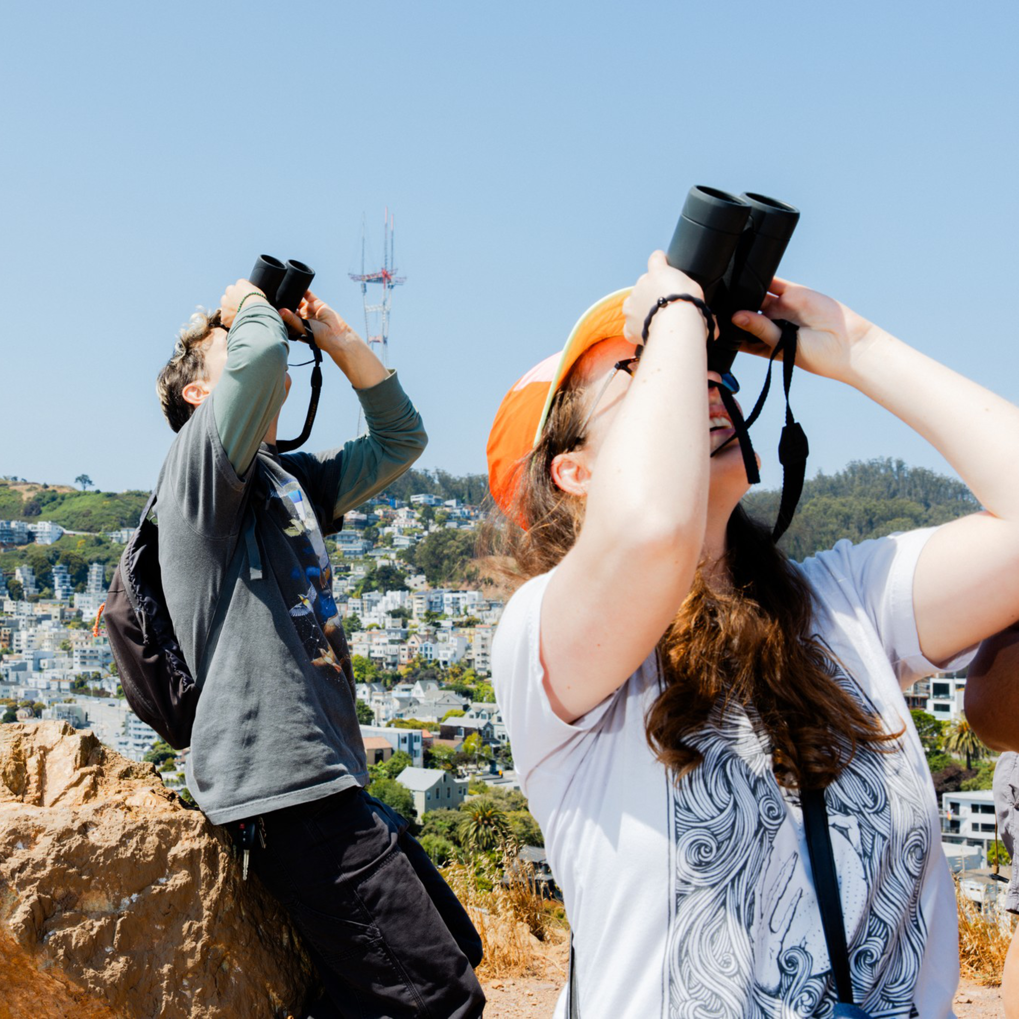 Two people stand on a rocky hilltop using binoculars, with a cityscape and greenery in the background. One wears an orange cap and the other a backpack.
