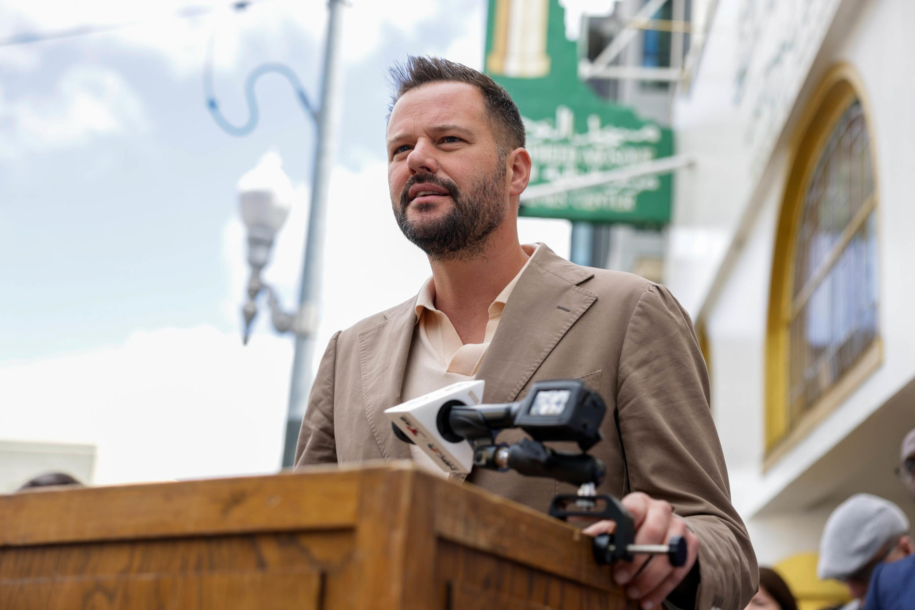 A man stands speaking at a podium with microphones, outdoors, daytime, with buildings and a lamp post in the background.