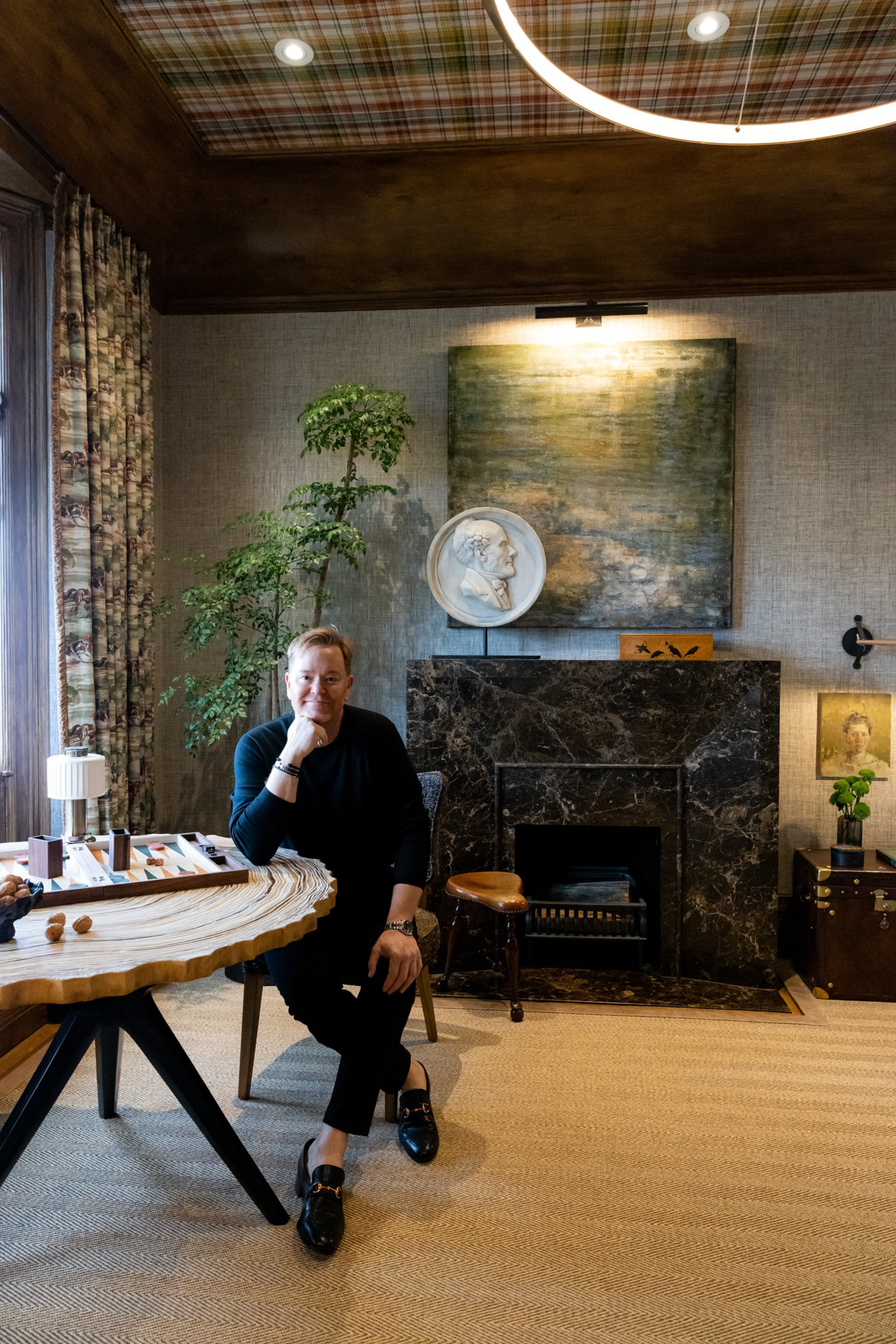 A man in black sits on a chair beside a wooden table in a cozy room featuring a black marble fireplace, a tall green plant, and artwork on the walls.