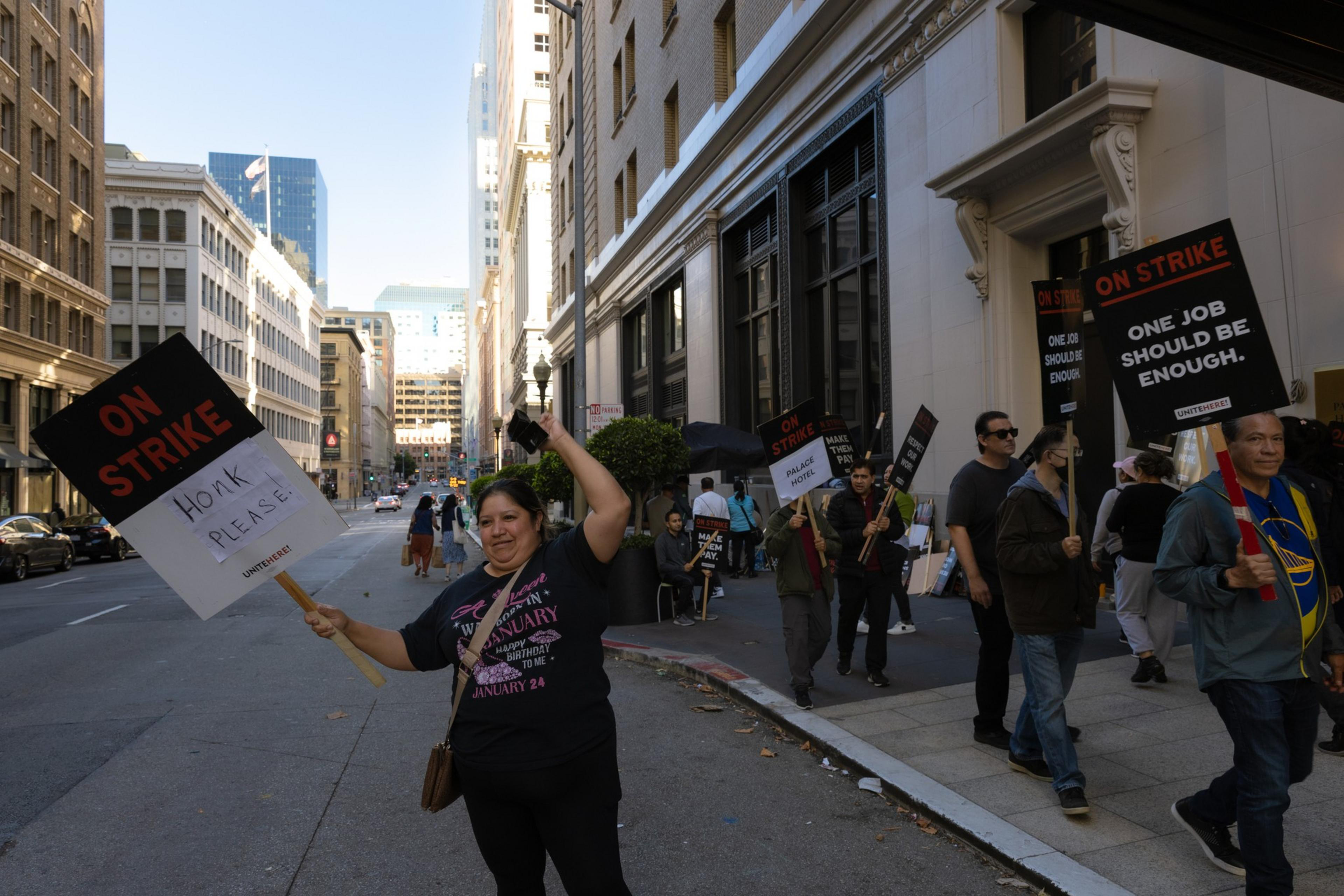 A group of people marches on a city street, holding strike signs with messages like &quot;On Strike,&quot; &quot;One job should be enough,&quot; and &quot;Honk please.&quot;