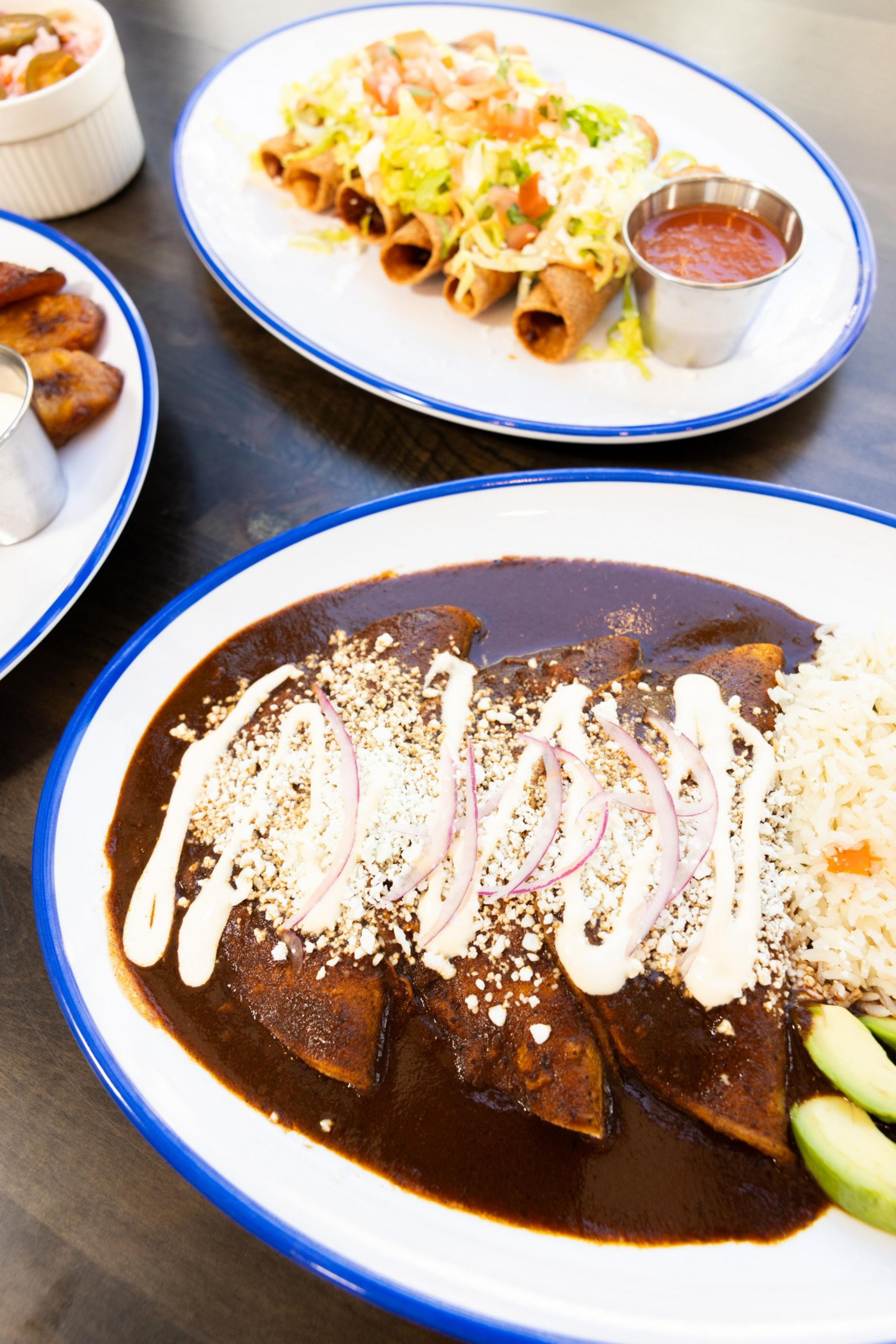 The image shows two oval plates of Mexican food, one with enchiladas covered in mole sauce, crema, and sliced onions, the other with crispy flautas topped with lettuce, tomato, and crema.