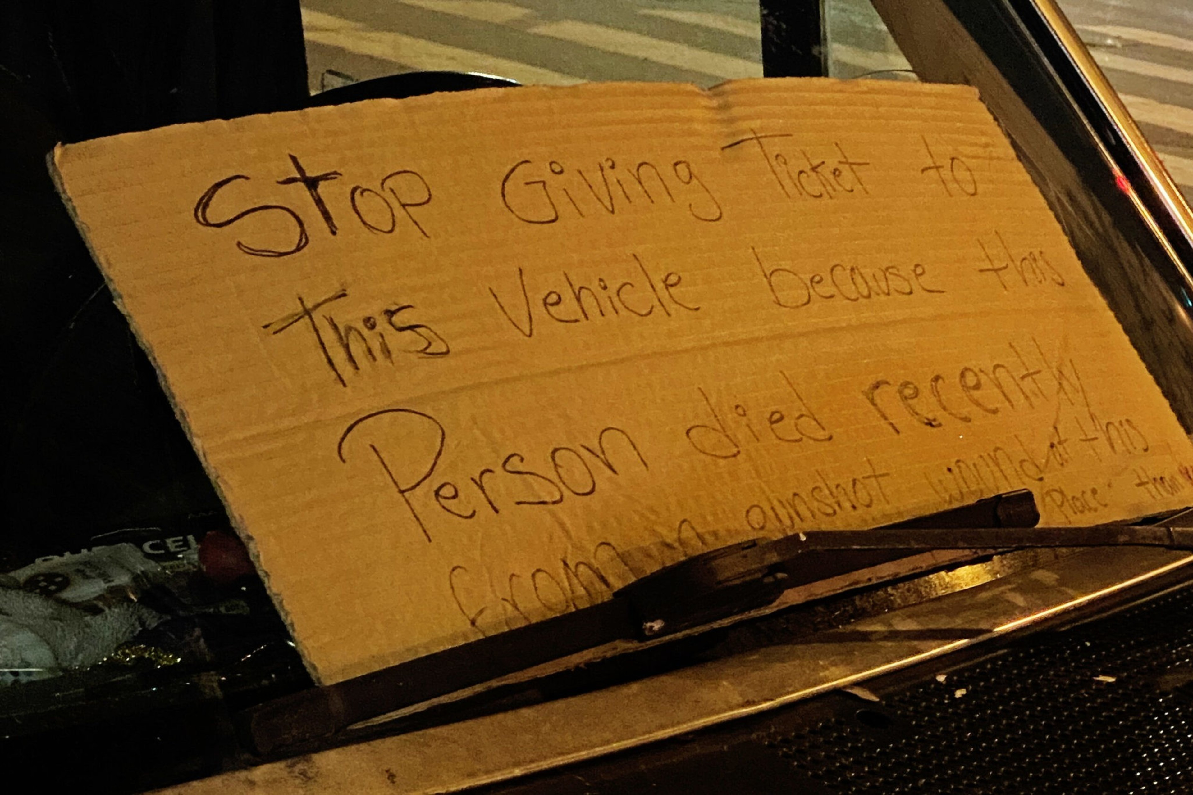 A handwritten cardboard sign on a car windshield pleads to stop giving tickets, explaining the owner recently died from a gunshot wound.