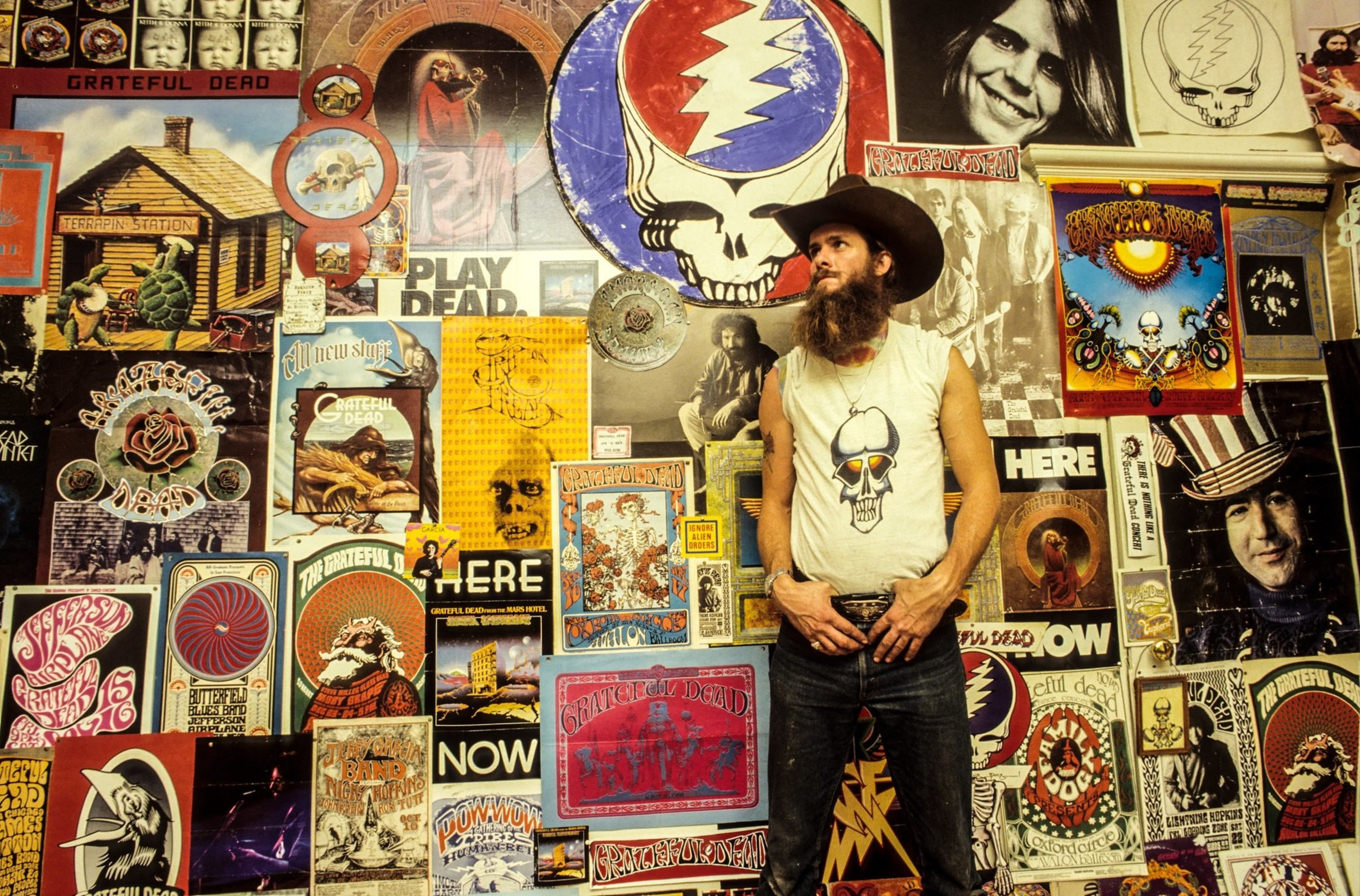 A bearded man in a cowboy hat and a skull t-shirt stands confidently in front of a colorful wall covered with various Grateful Dead posters and artwork.