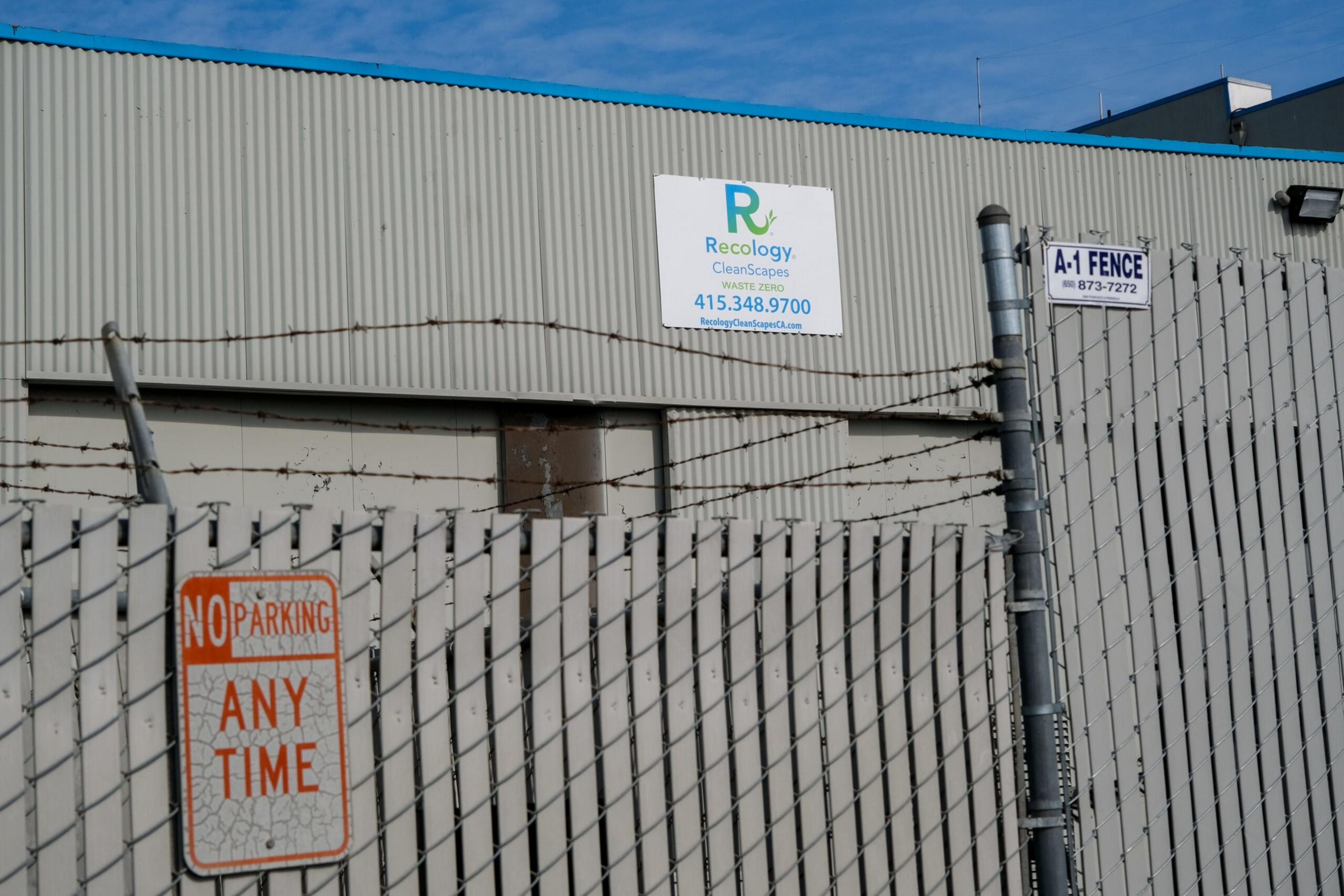 A barbed wire fence with "No Parking Any Time" and "A-1 Fence" signs stands in front of a beige building with a Recology Cleanscapes sign.