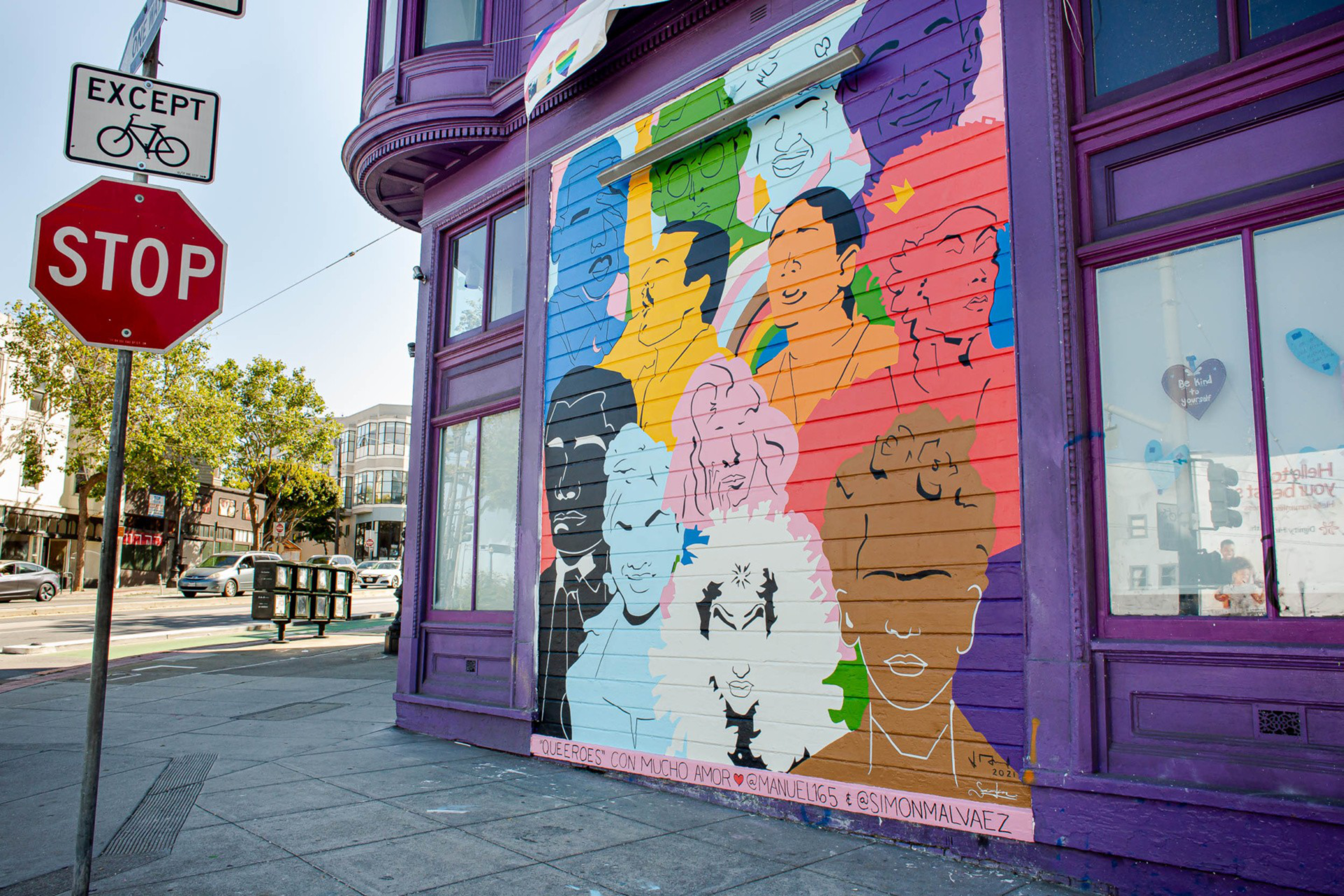 A colorful mural on a purple building wall depicts various outlined faces in multiple bright colors next to a stop sign with a bicycle exception.