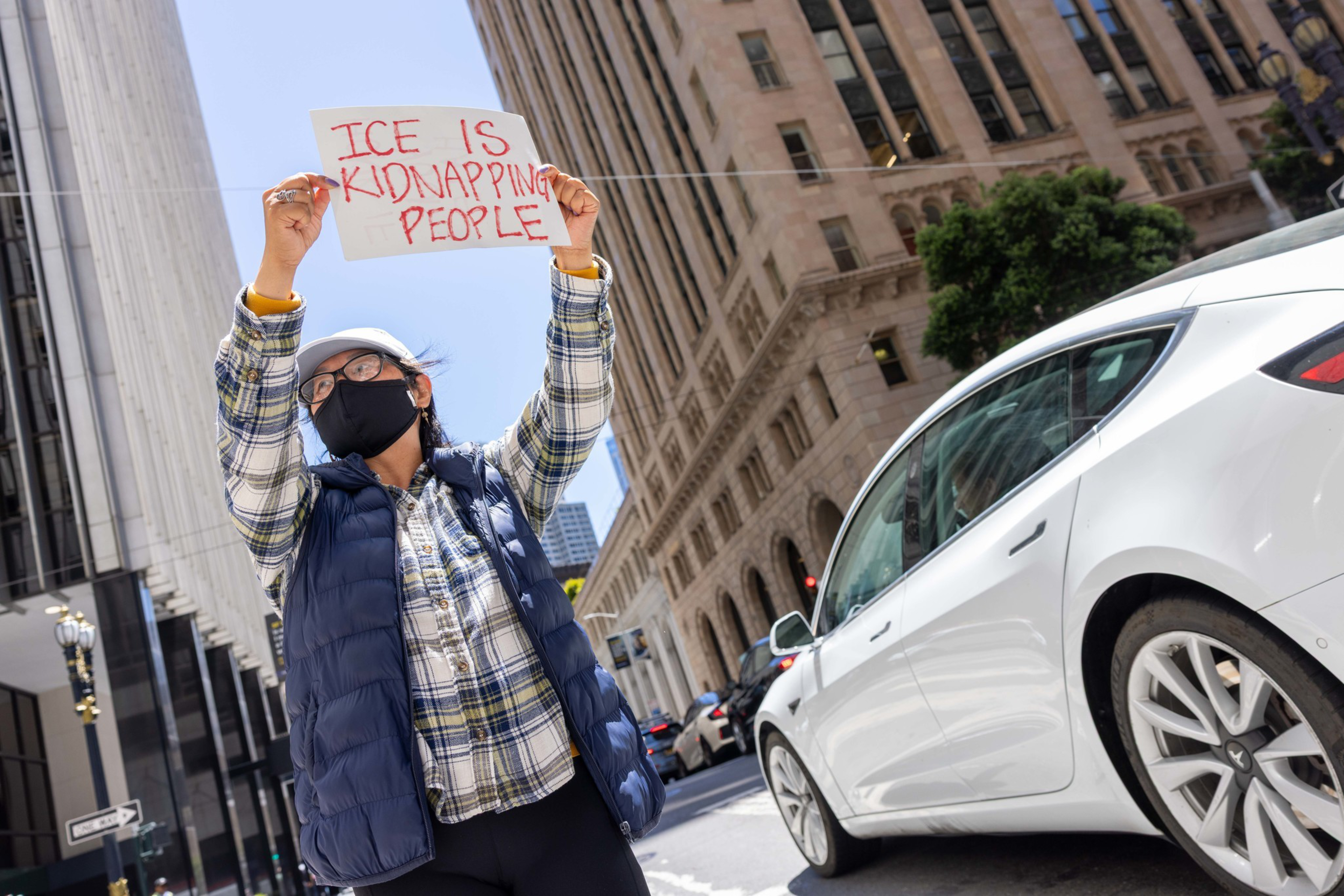 A person wearing a mask holds a sign saying "ICE IS KIDNAPPING PEOPLE" on a city street, with tall buildings and a white car in the background.
