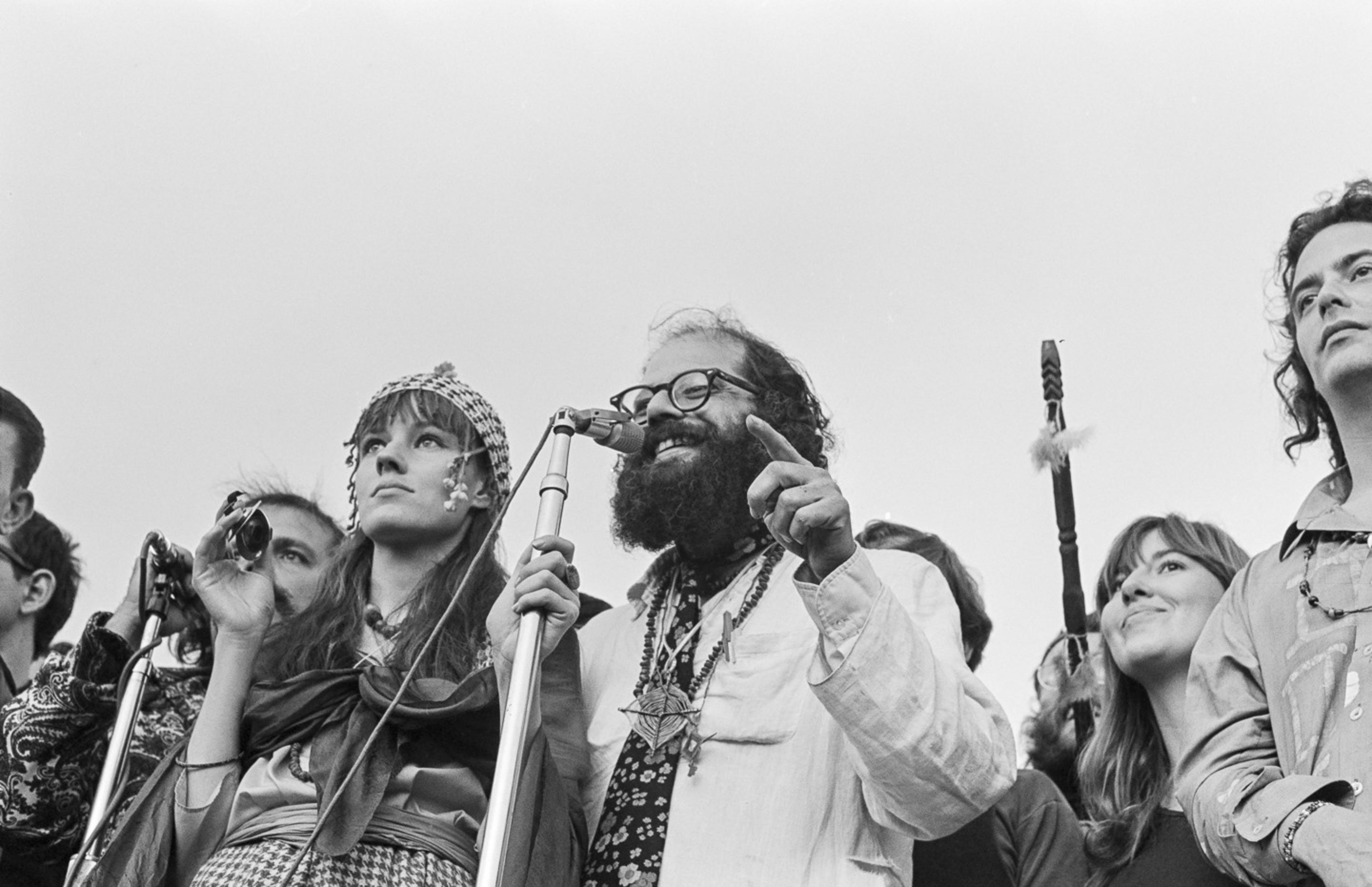 A group of people stands together, with a man speaking into a microphone. They're dressed in 1960s style, with beads and vibrant clothing, looking cheerful and expressive.