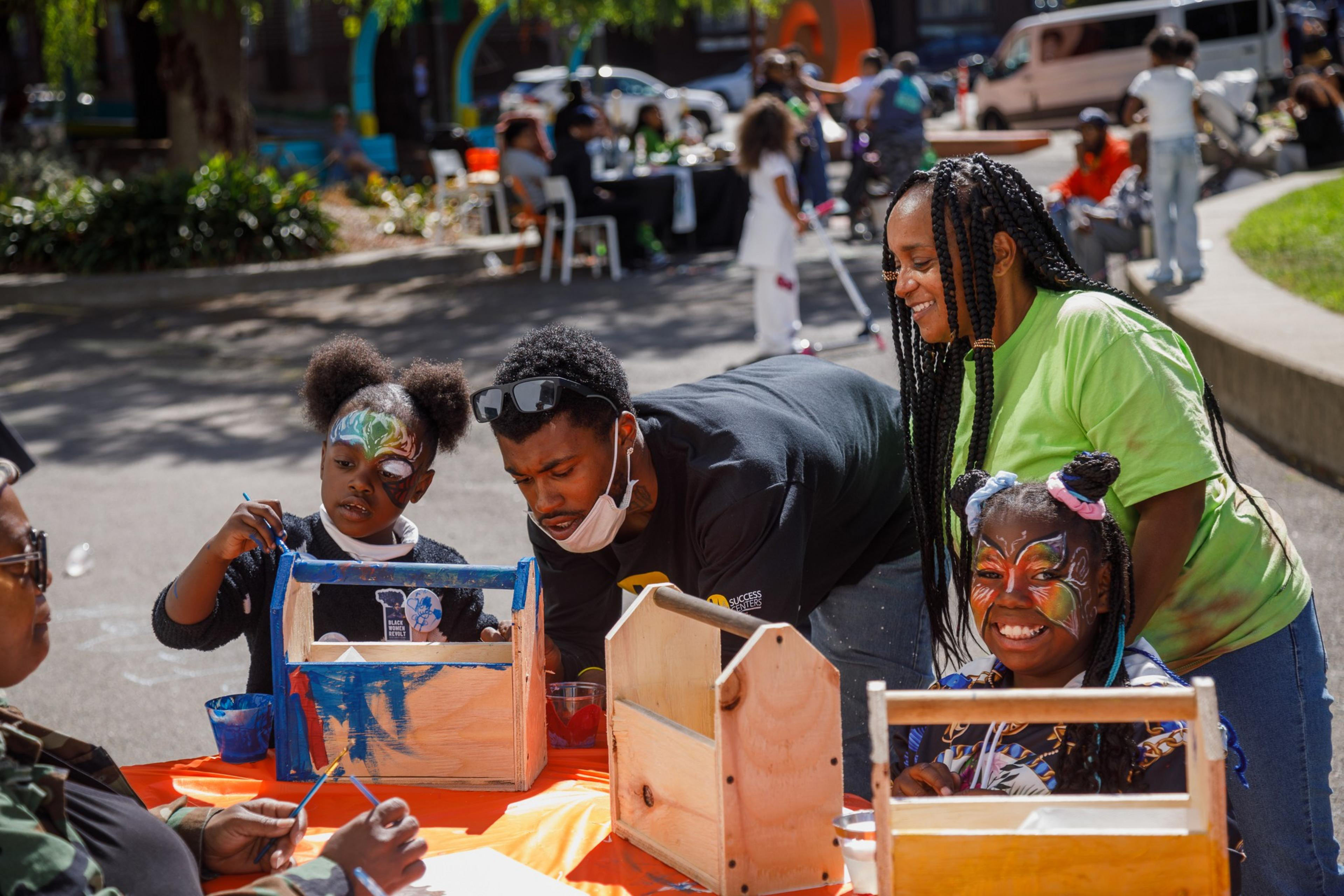A family is engaged in crafts, with two children showing face paint, at an outdoor event with other attendees.