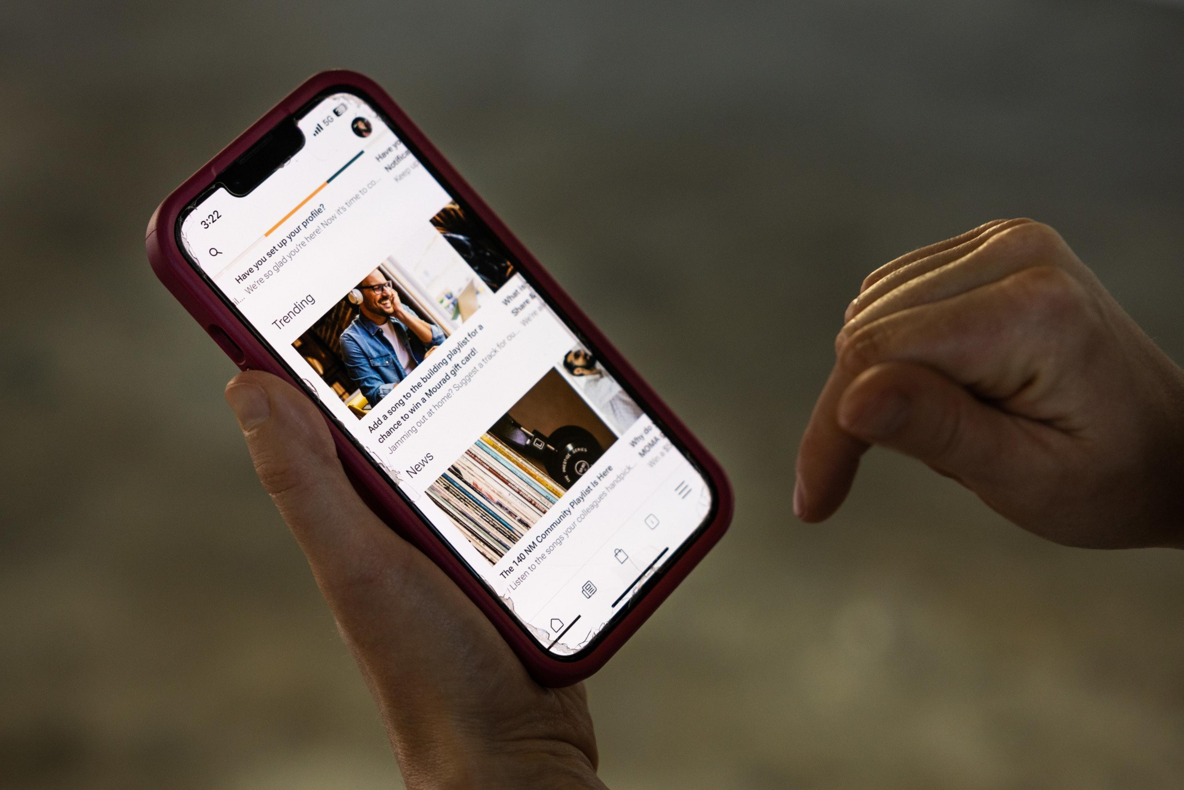A smartphone, held in a pink case, displays a news feed with articles. A hand next to it appears to be gesturing, indicating interaction with the content.