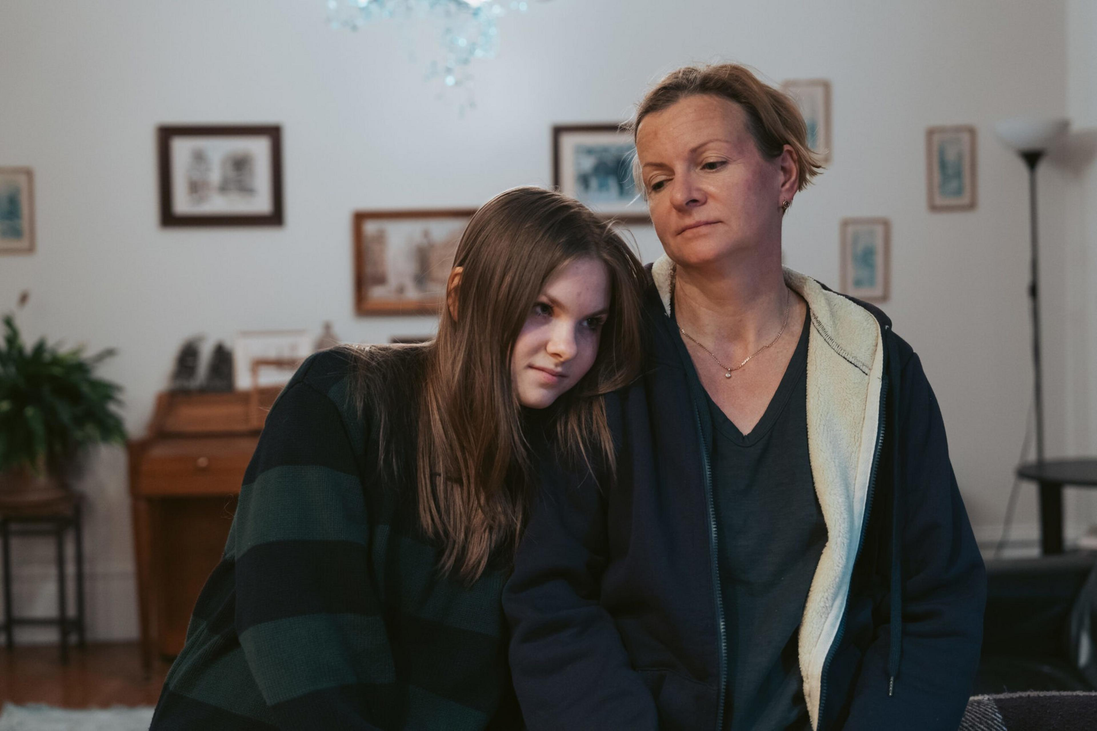 A teenage girl leans her head on an older woman's shoulder while both sit indoors, looking thoughtful and somber.