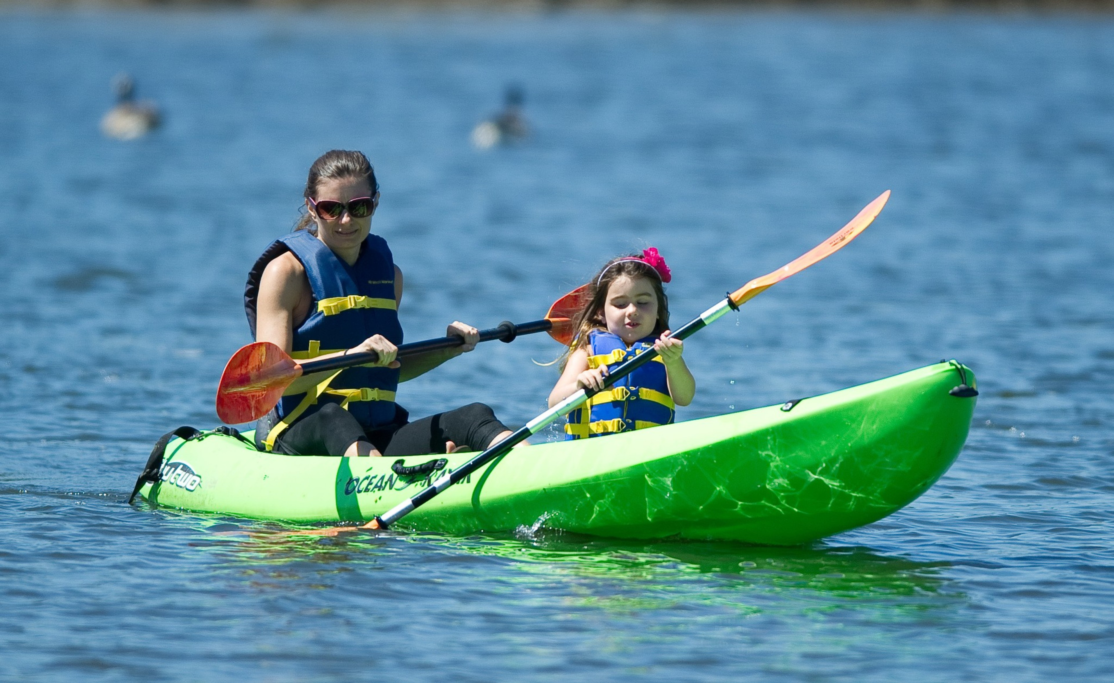 Two people in life jackets are paddling a green kayak on calm blue water.