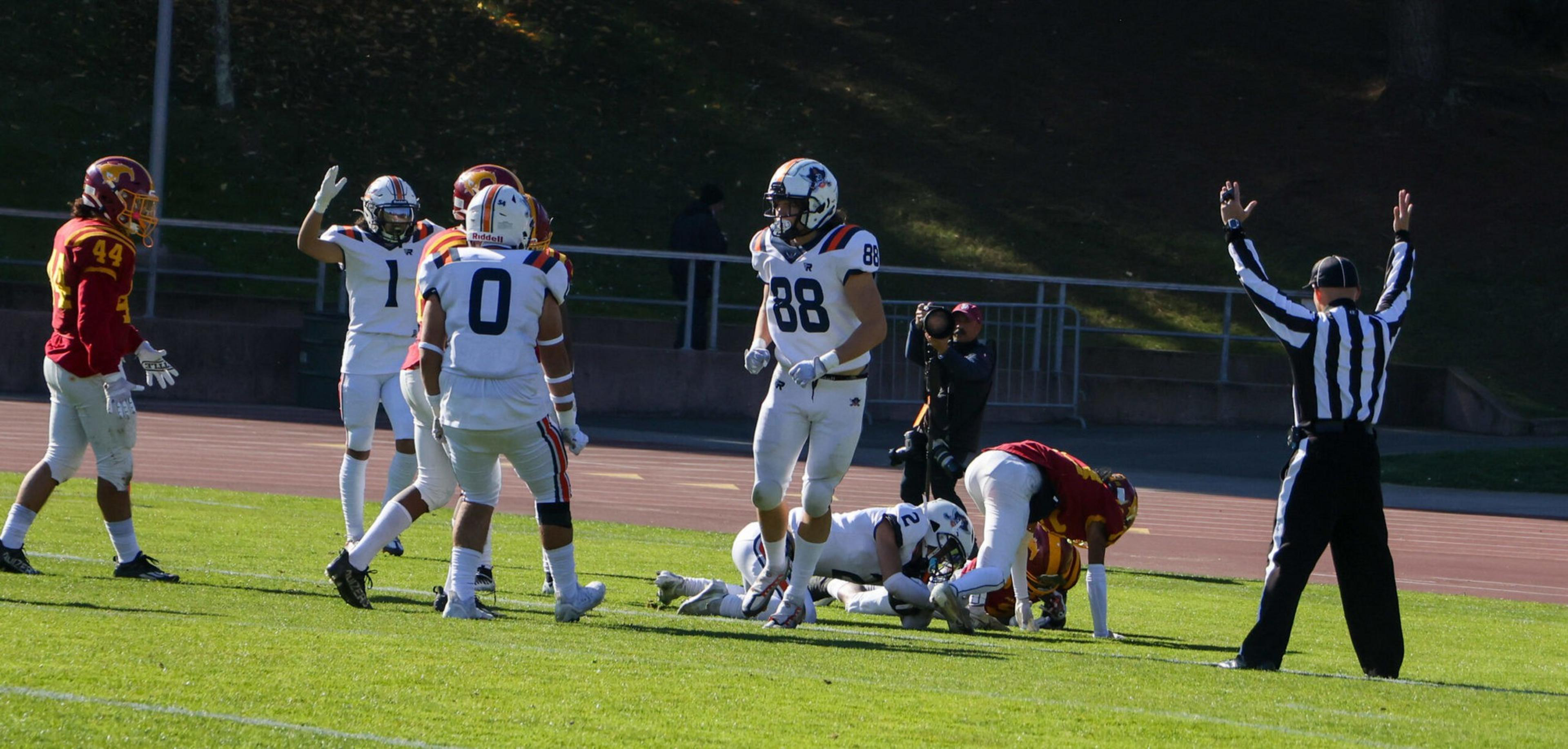 Football players in white celebrate a touchdown while a player in red lies on the ground holding the ball, and a referee signals a score with raised arms.