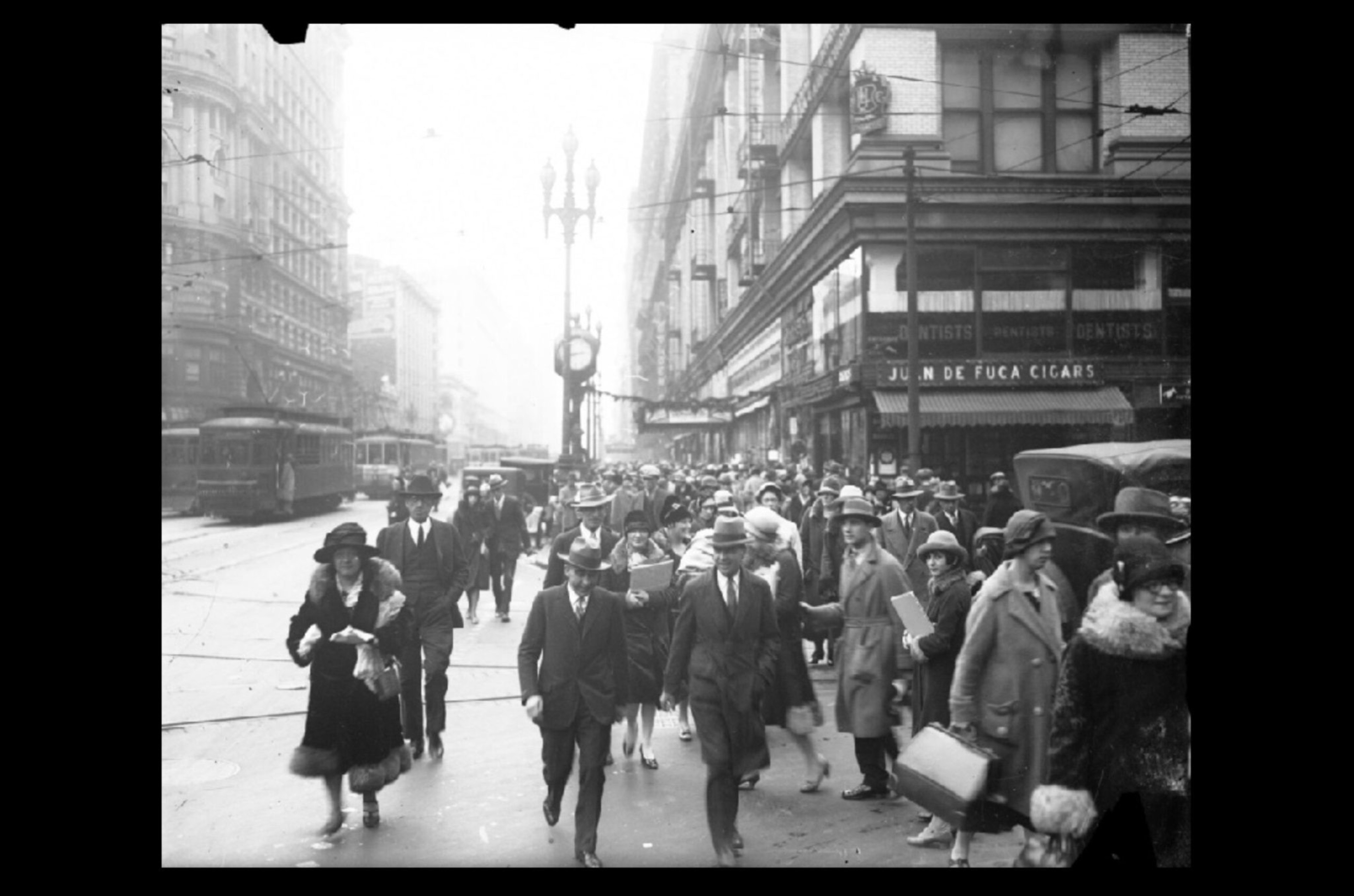 Busy city street filled with men and women in early 20th-century attire, walking past storefronts including one labeled "Juan de Fuca Cigars."